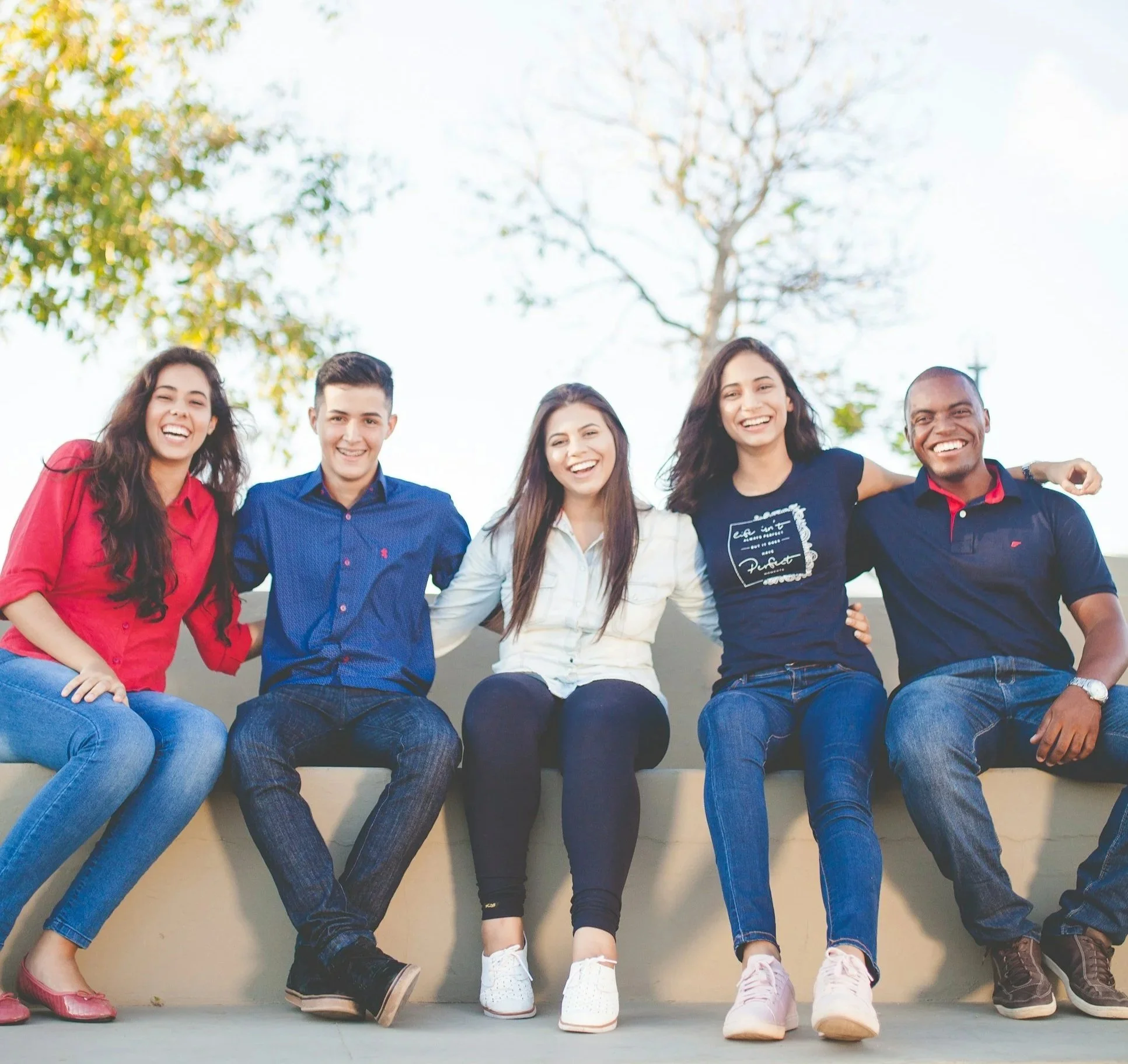 Group of five diverse young adults sitting outdoors on a wall, smiling and enjoying themselves.