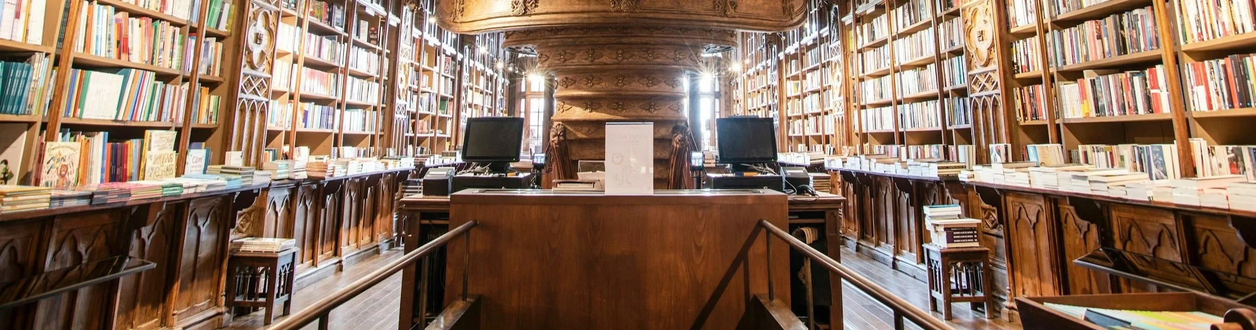 Interior of a large wooden library with high shelves filled with books, a wooden desk with two computer monitors, and sunlight streaming through windows.