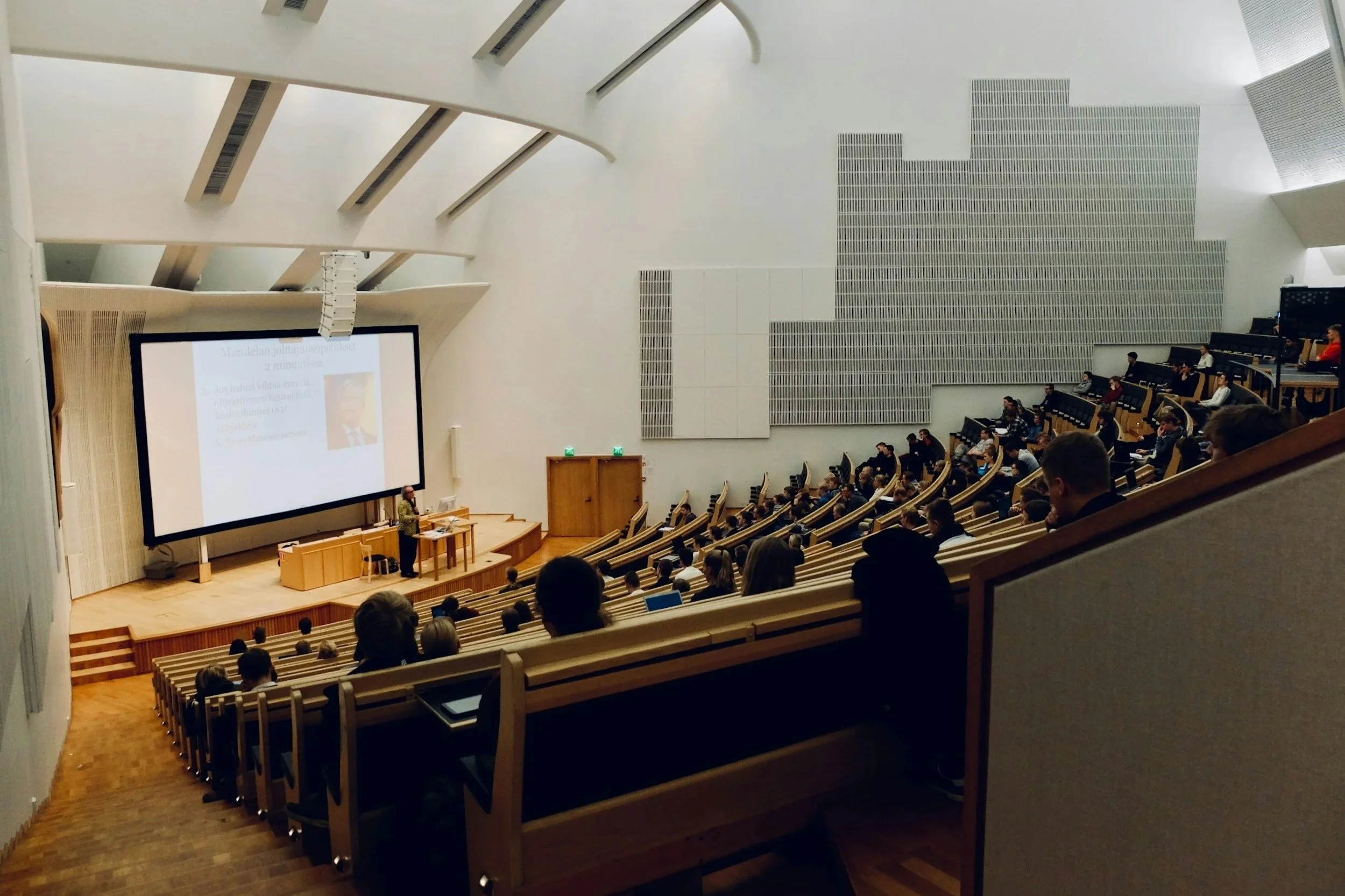 A lecture hall with tiered seating filled with people, a speaker at a podium in front of a large screen displaying a presentation, and a white interior with acoustic panels on the wall.