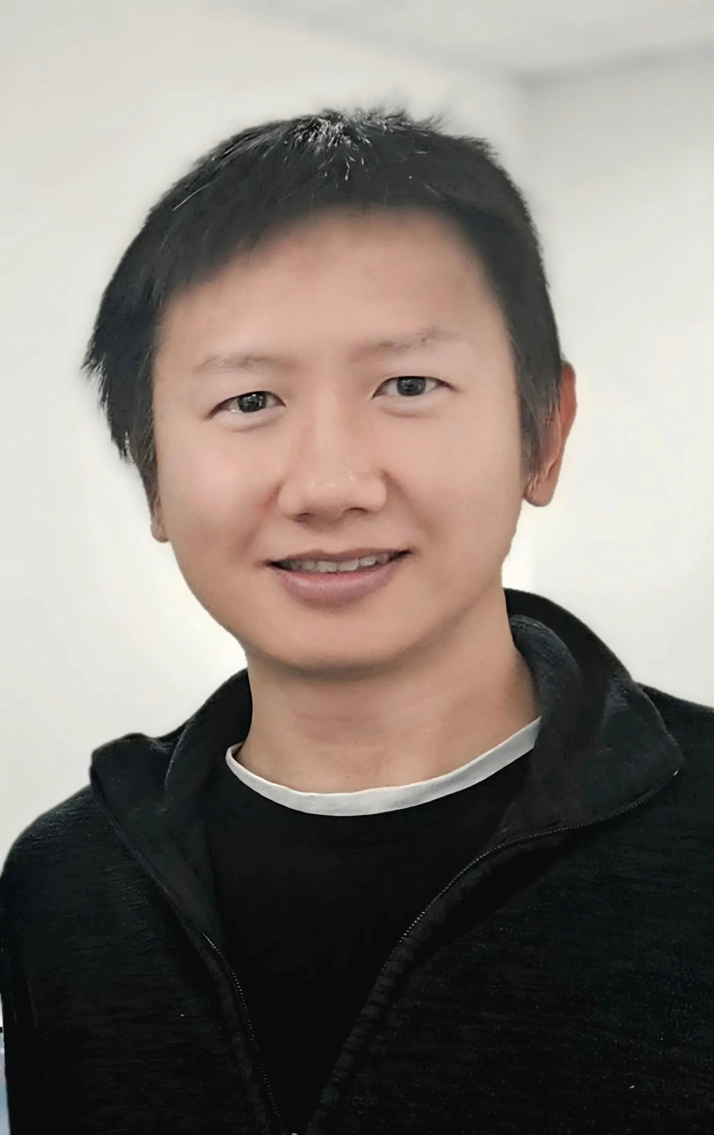 A young man with short black hair and light skin, smiling, wearing a black zip-up jacket with a white shirt underneath, posed against a plain light-colored background.