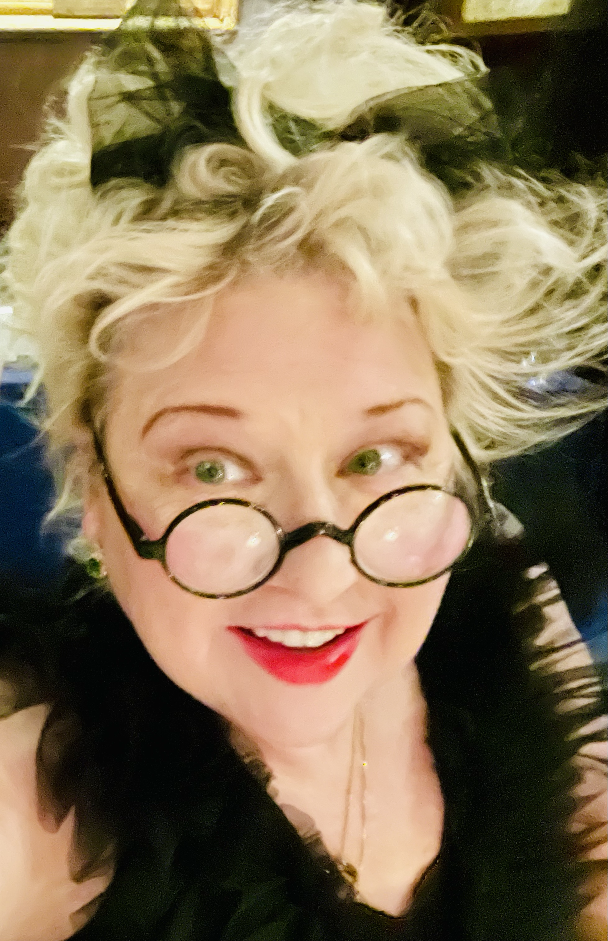 A woman with curly blonde hair wearing round glasses with black frames, red lipstick, and a black top, smiling and looking up.