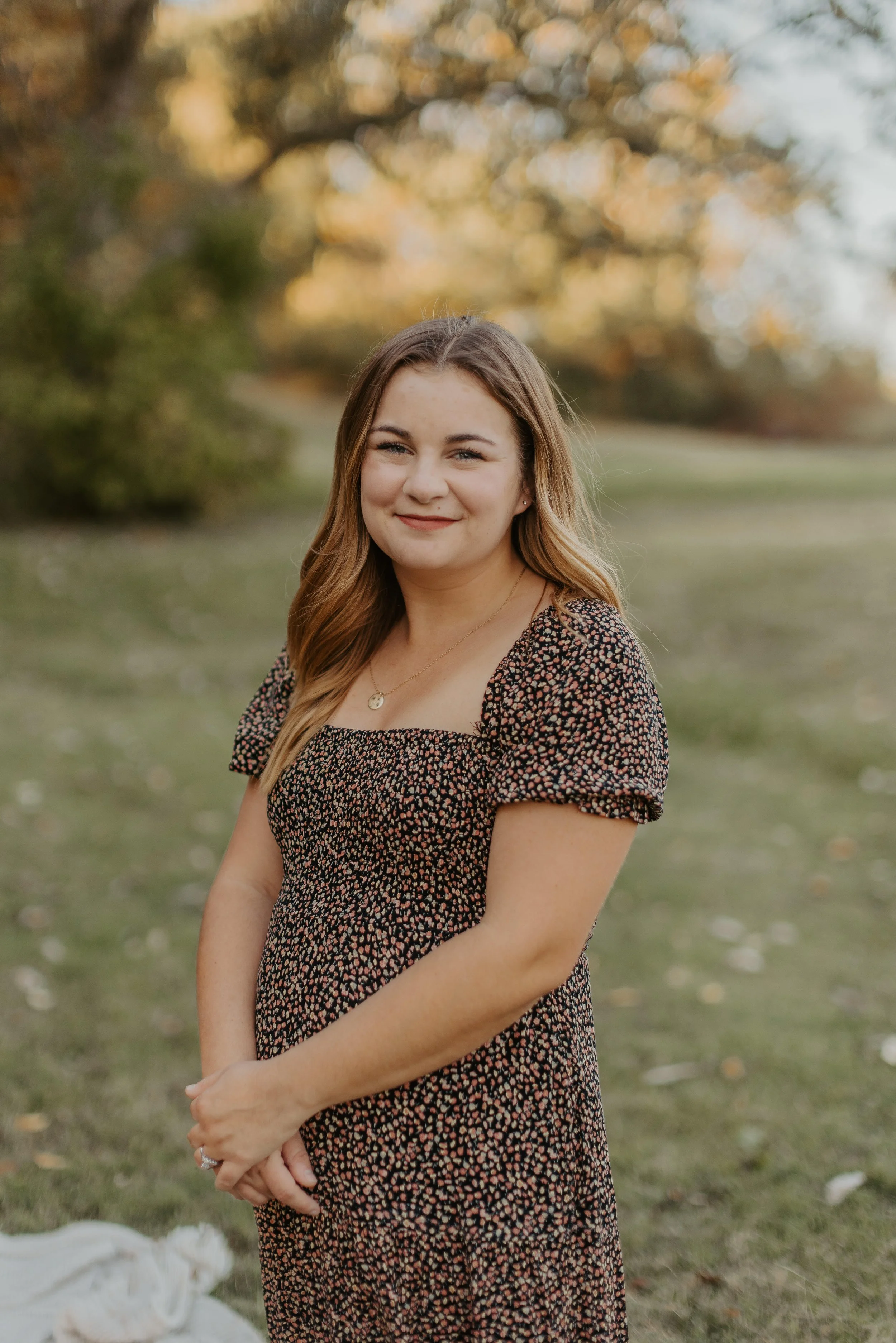 A young woman with long, wavy hair standing outdoors in a park, wearing a black dress with small floral print, smiling and facing the camera with hands clasped in front of her.