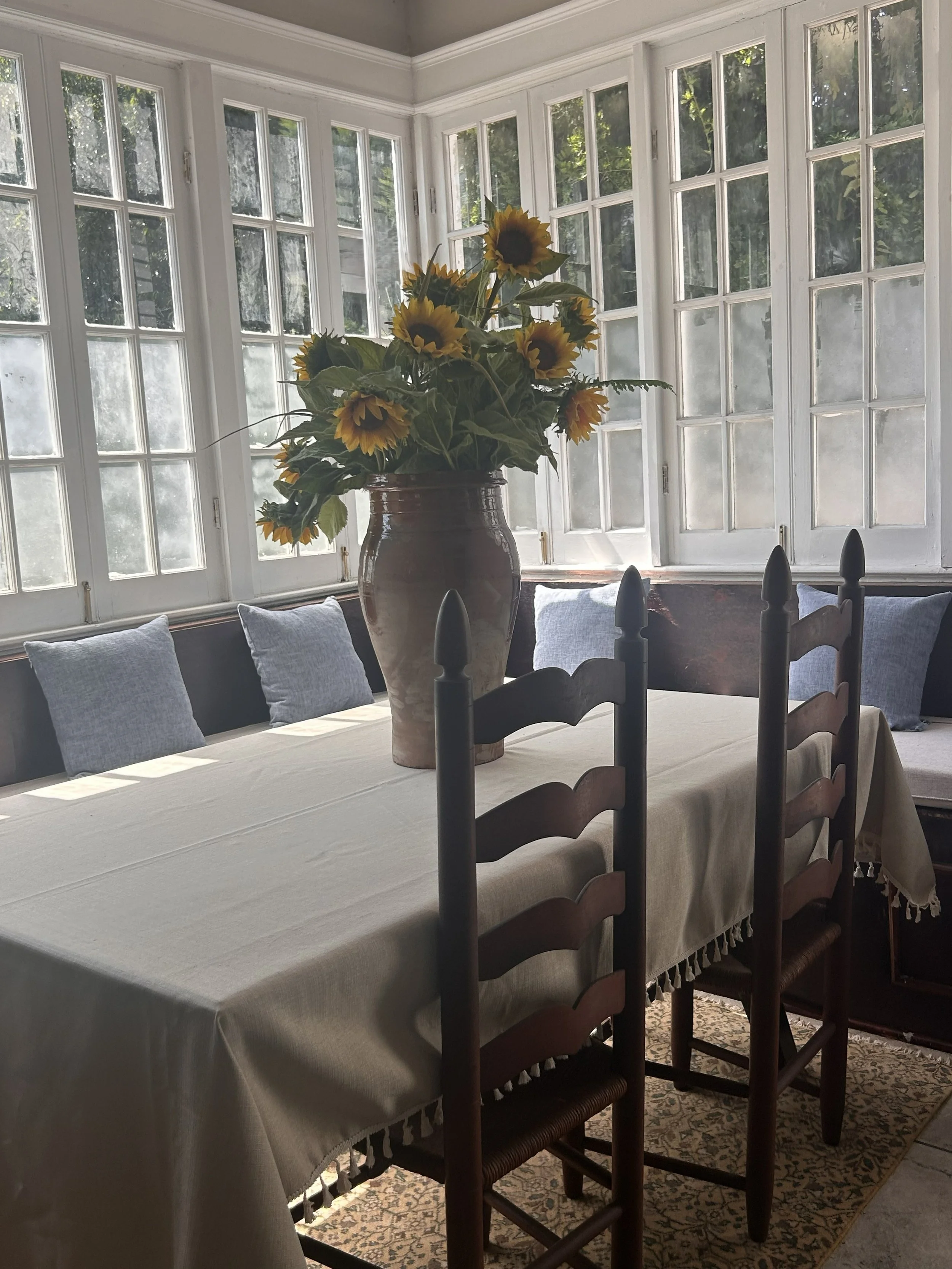 A dining room with a long rectangular table covered with a cream-colored tablecloth. On the table, there is a large vase with a bouquet of sunflowers. The background features a large bay window with multiple panes, letting in natural light and showing greenery outside. There are several light-colored cushions on a built-in bench along the wall and a patterned area rug under the table.
