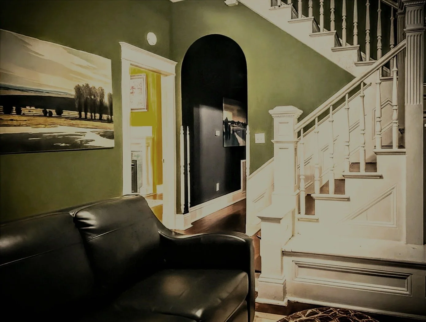 Interior view of a living room with a black leather sofa, green wall with a landscape painting, a staircase with white wood railing, and a dark passageway leading to another room.
