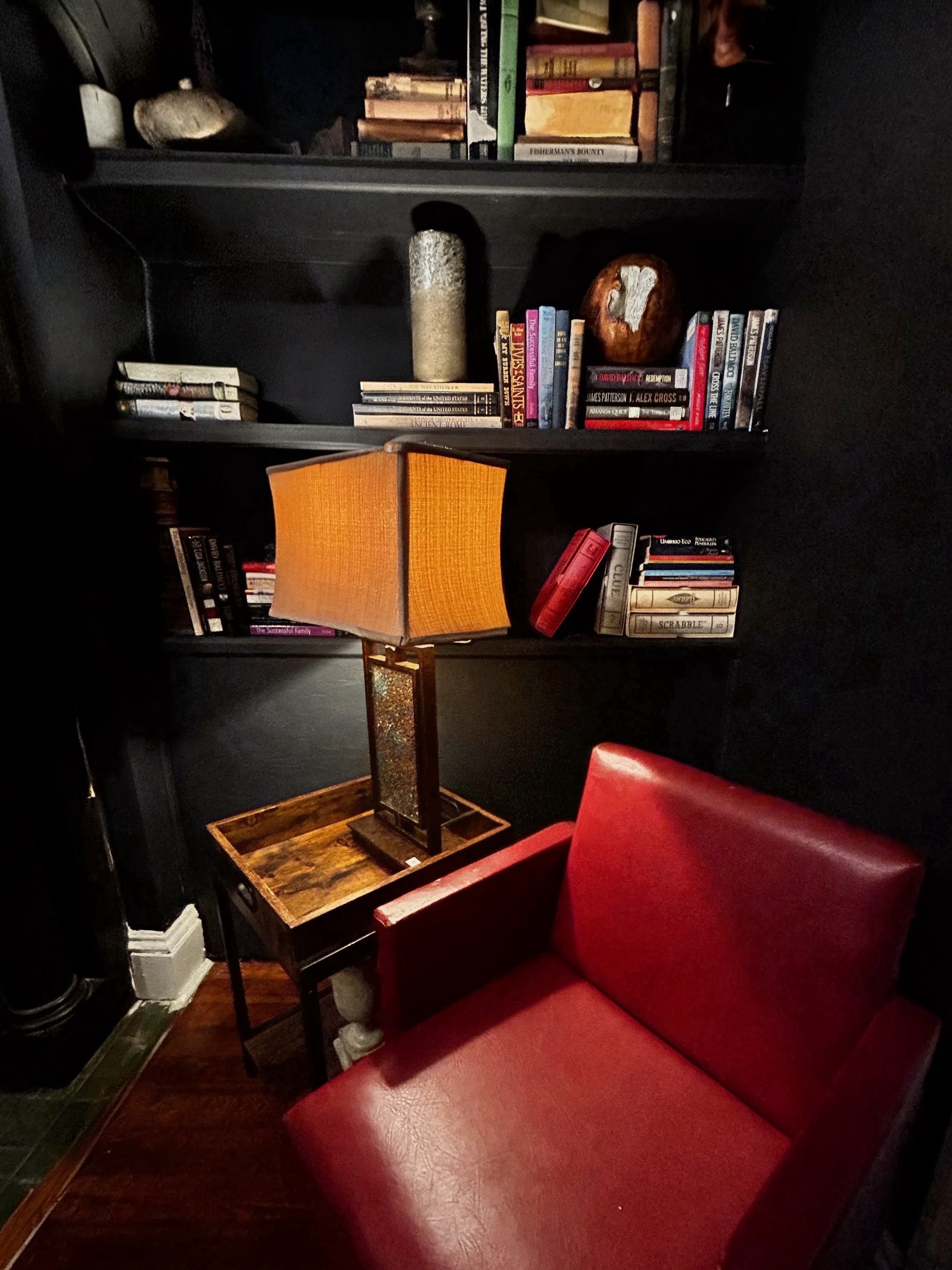 A cozy corner with a red leather armchair, a wooden side table with a lamp, and black bookshelves filled with books, decorative objects, and small sculptures.