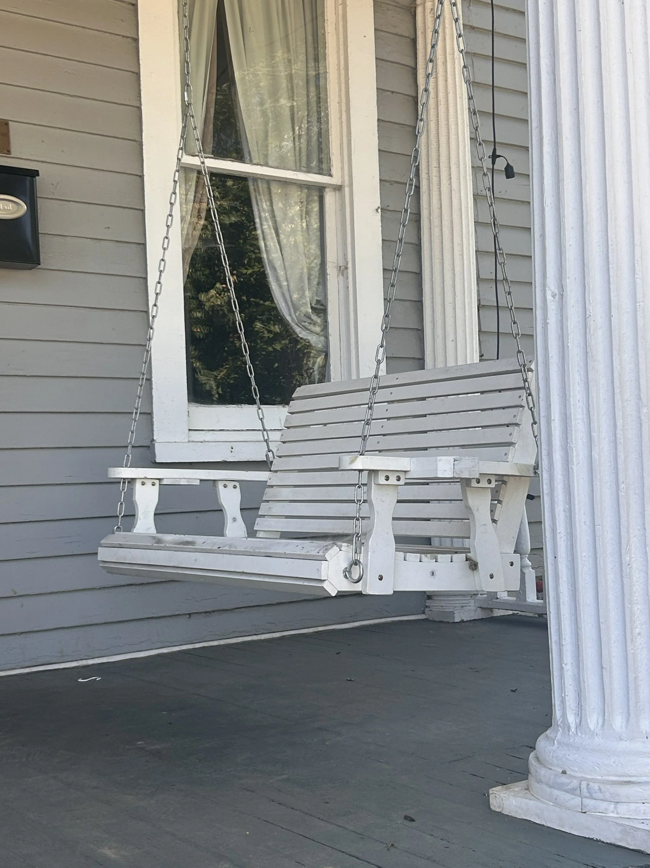 White porch swing with chained support hanging in front of a house with light gray wooden siding and window with curtains, next to a white column.