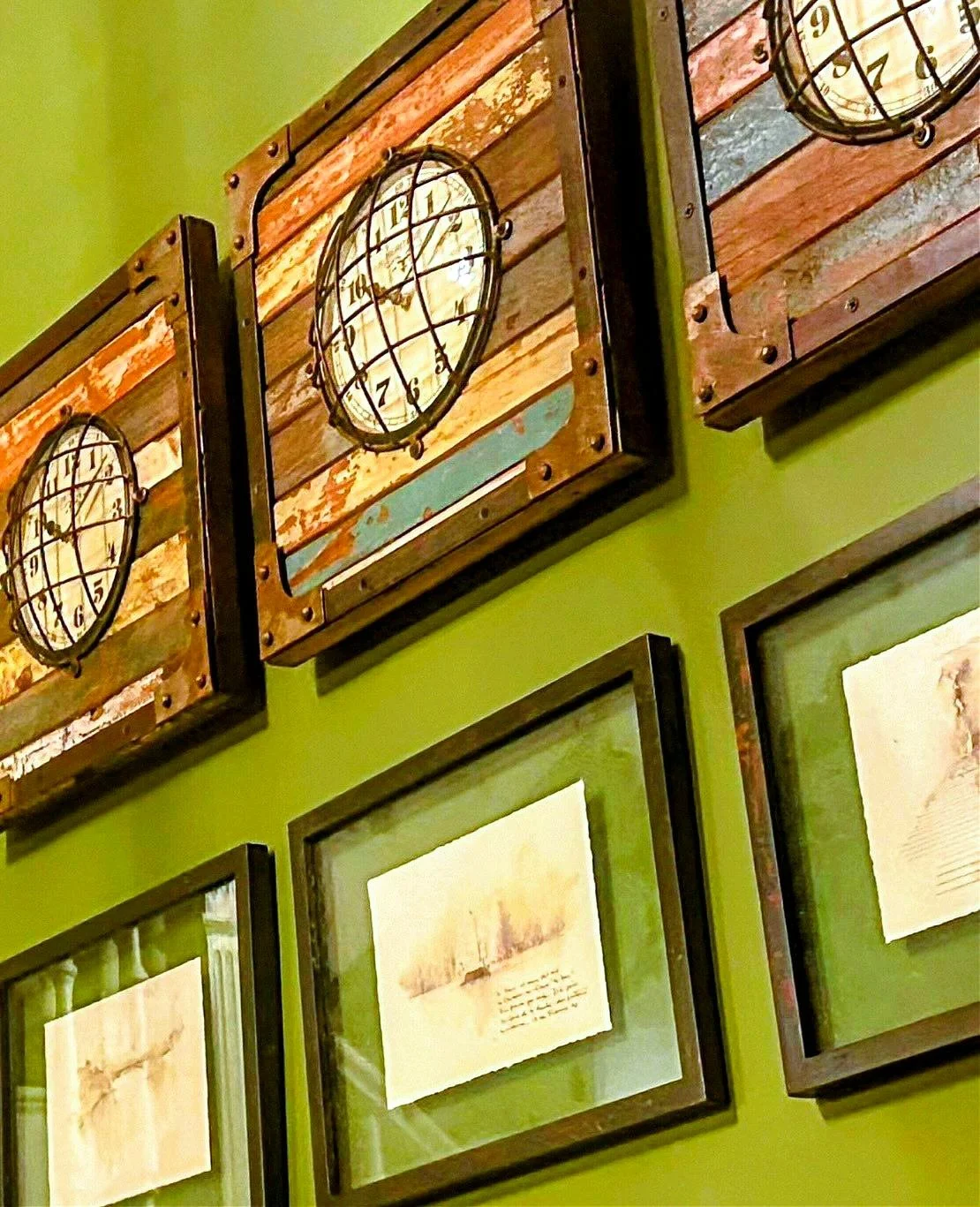 Decorative wall clocks with a rustic, vintage design mounted on a green wall, accompanied by framed artwork.