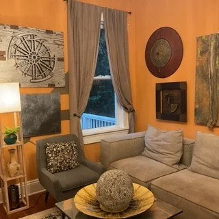 Living room with a light gray sofa and a gray armchair, wooden side table with a lamp and plant, window with curtains, orange walls decorated with art and a mirror, coffee table with a large decorative ball.