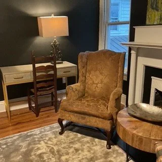 An interior room with a brown upholstered armchair, a wooden side table with a metallic handle, a white fireplace, a window with blinds, a desk with a chair and a lamp, and a patterned rug on hardwood floor.
