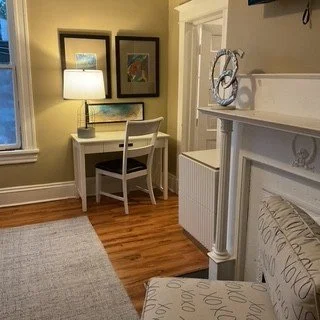 A cozy corner of a room with a white desk, a white chair, and a lamp, decorated with framed artwork on the wall.