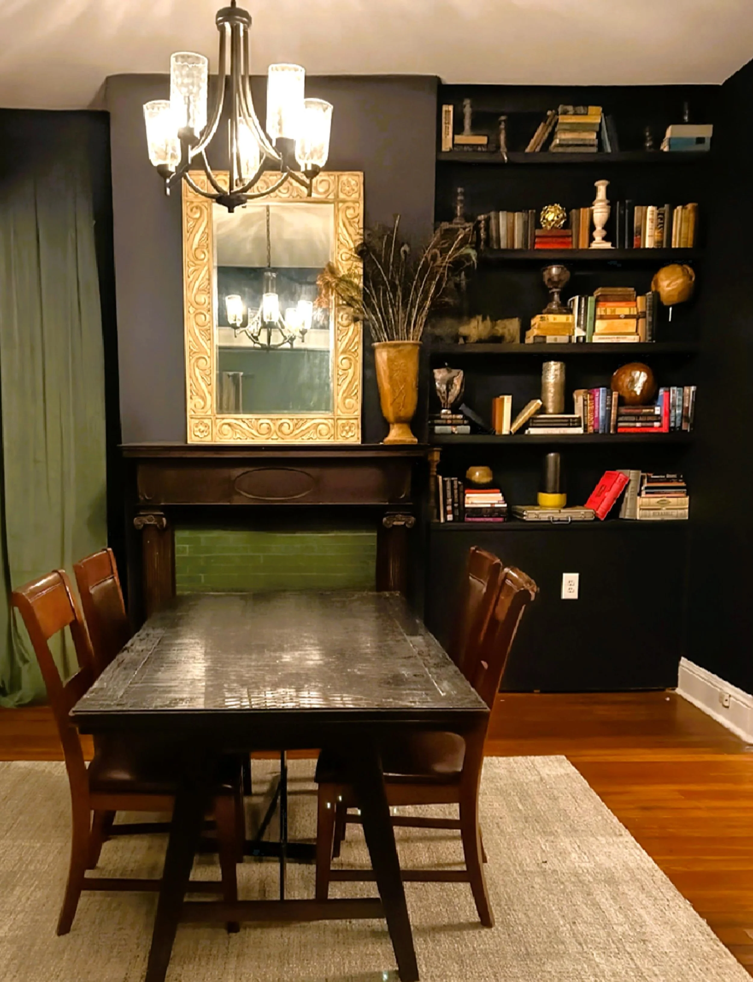 A study with a rectangular table and four wooden chairs, a large ornate mirror on the wall, a vintage chandelier hanging from the ceiling, and a black bookshelf filled with books and decorative items in the background.