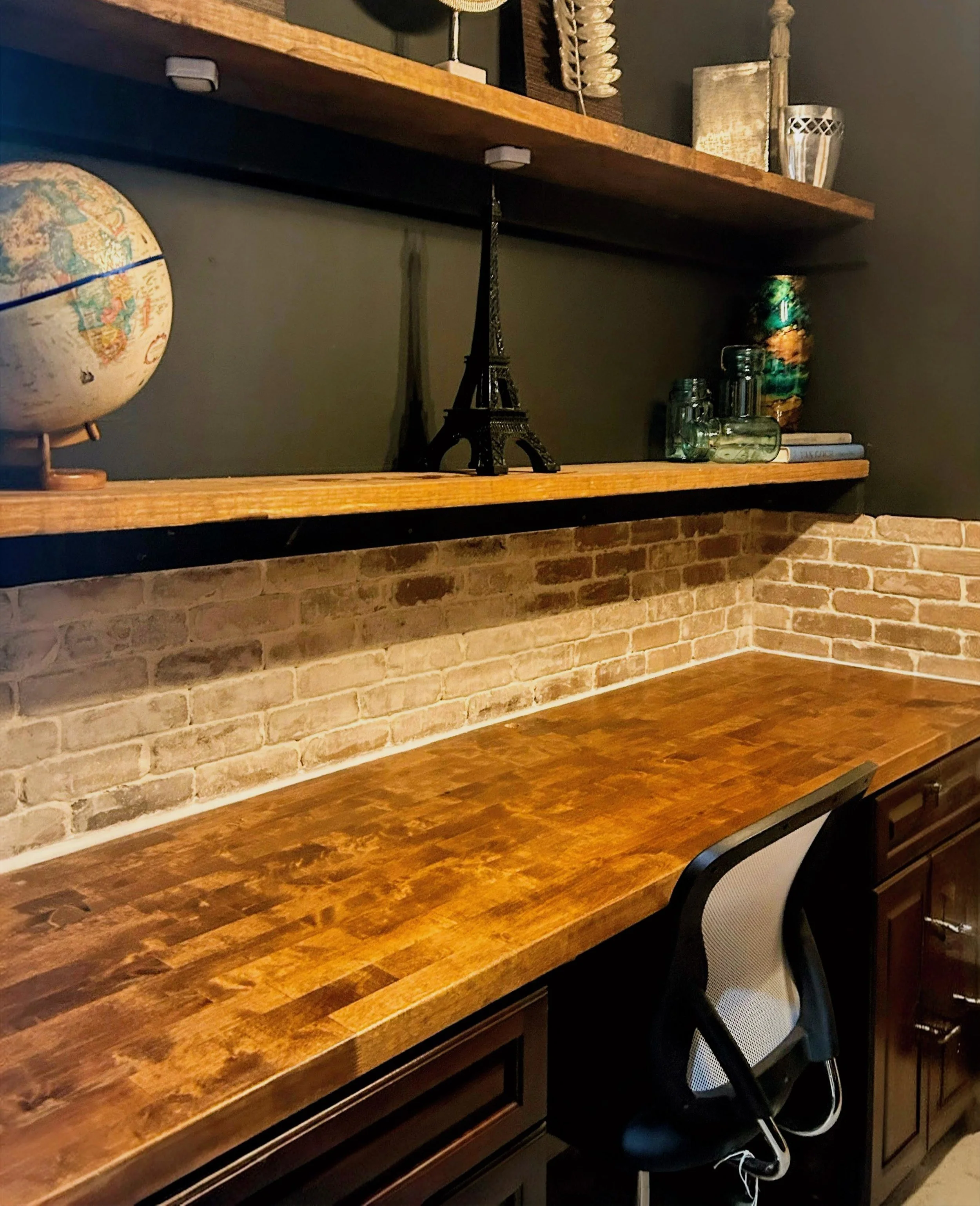 A room with a wooden desk and a black mesh office chair. The wall behind features a brick pattern. On the wooden shelf above, there is a globe on the left, a small Eiffel Tower figurine in the center, and decorative vases and books on the right.