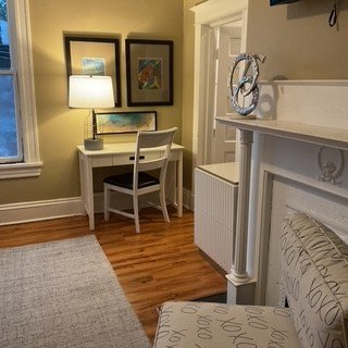 A cozy corner with a small white desk, a white chair, and three framed pictures on the wall. There is a white lamp on the desk and natural light from a window. A white cabinet and a decorative mirror are on the adjacent wall.