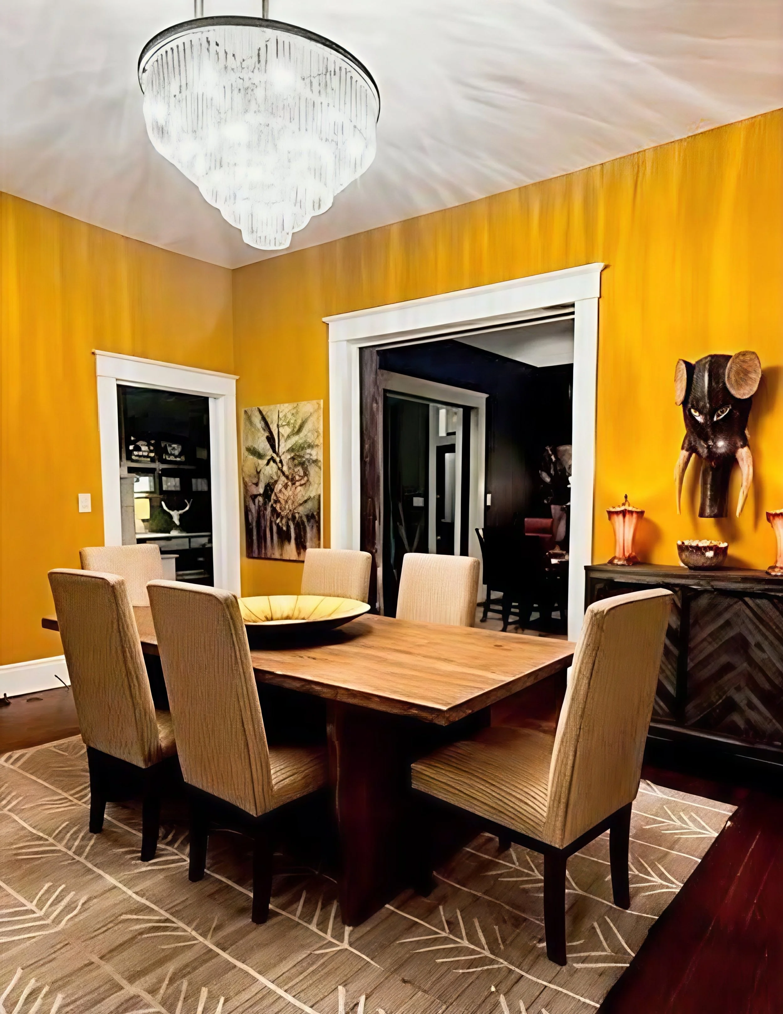 Dining room with a wooden table surrounded by six beige upholstered chairs, a chandelier on the ceiling, yellow walls, a painting on the wall, and a side table with decorative items.