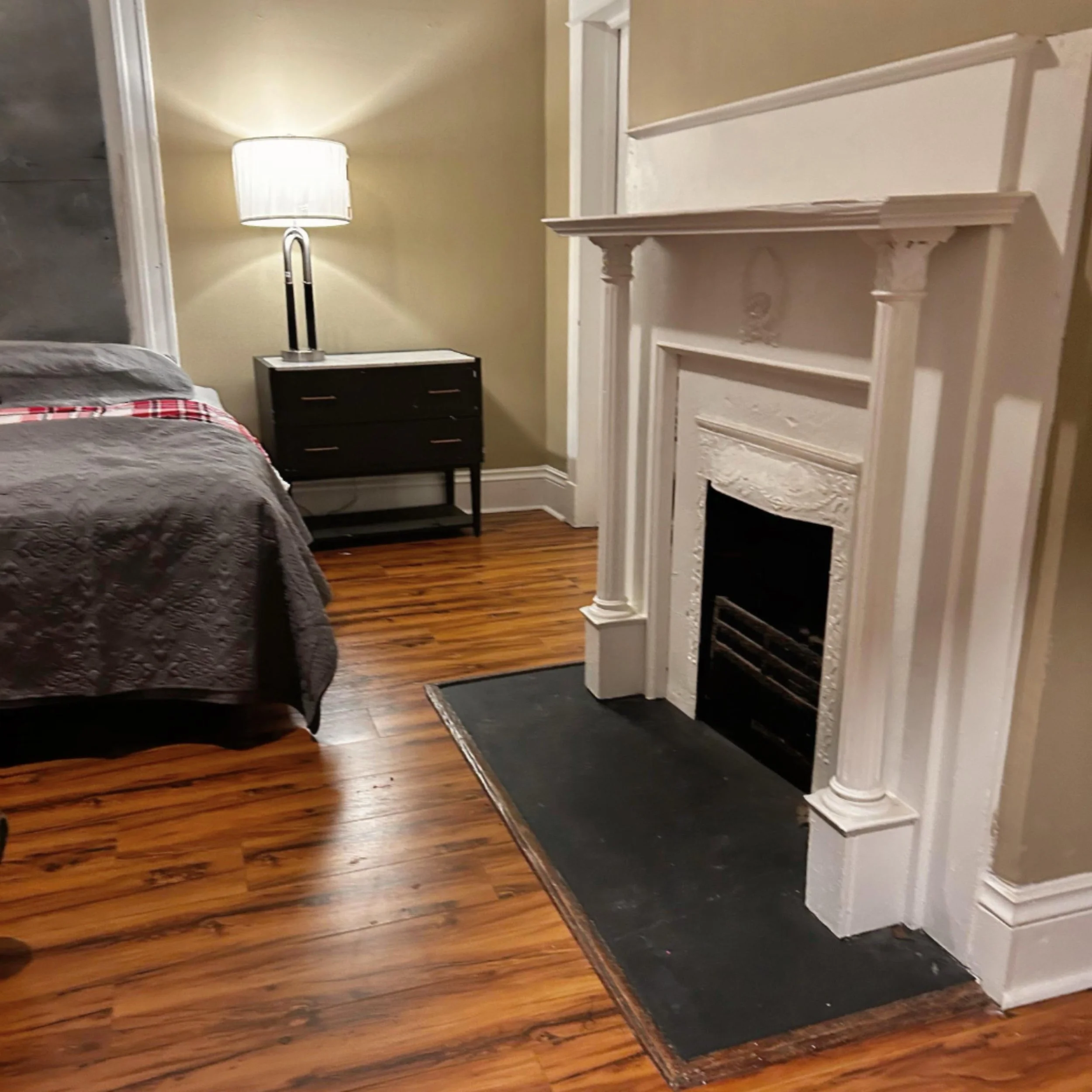 A bedroom featuring a bed with a gray quilt and a red plaid blanket, a black nightstand with a lamp, a white decorative fireplace, and hardwood floors.