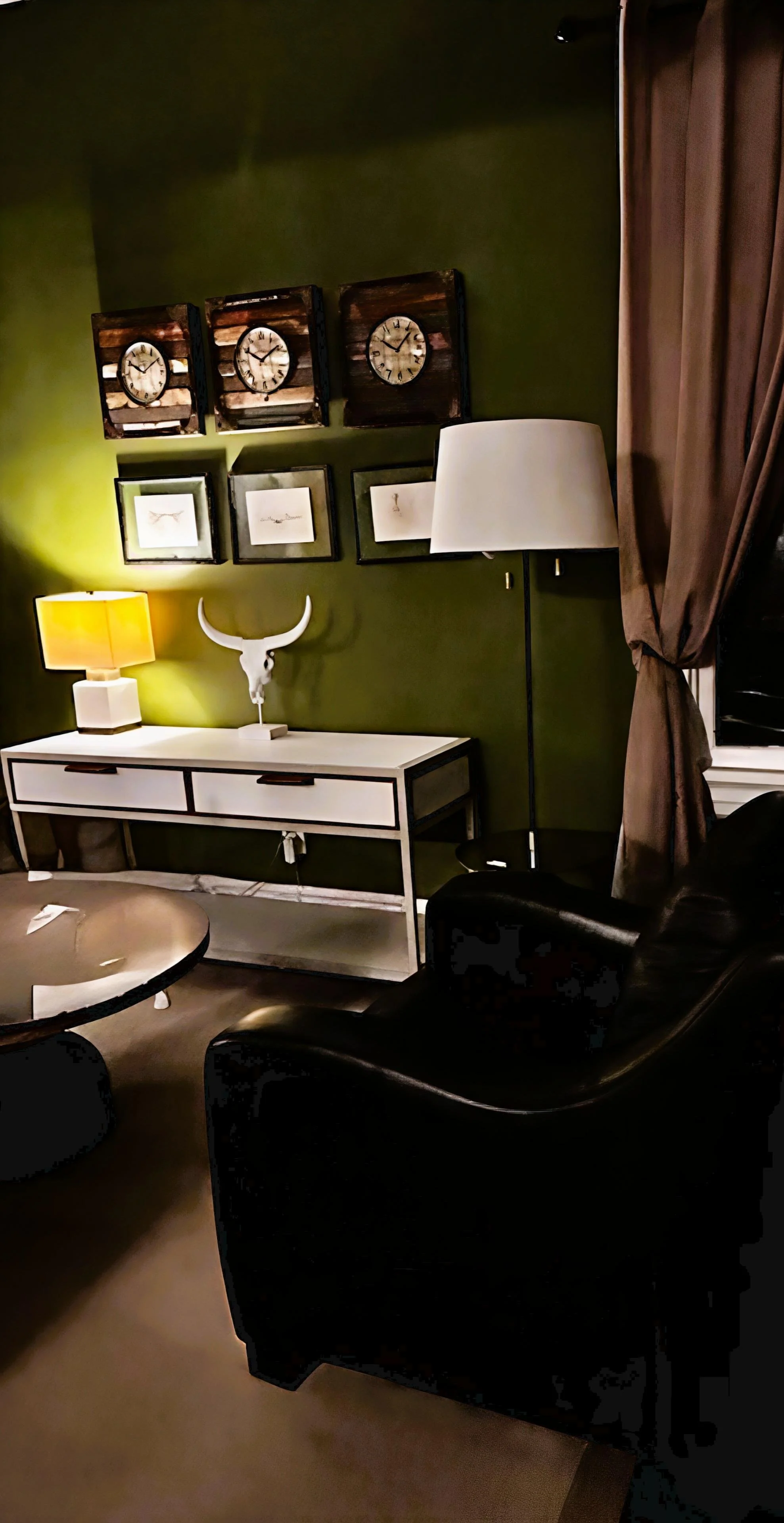 Living room with dark green wall, wall art including clocks and drawings, white console table with a lamp, decorative horn skull, black armchair, beige curtains, and a small round table.