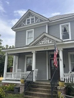 A two-story house with gray siding and white trim, featuring a porch with steps, an American flag hanging, and a decorative gable above the entrance.