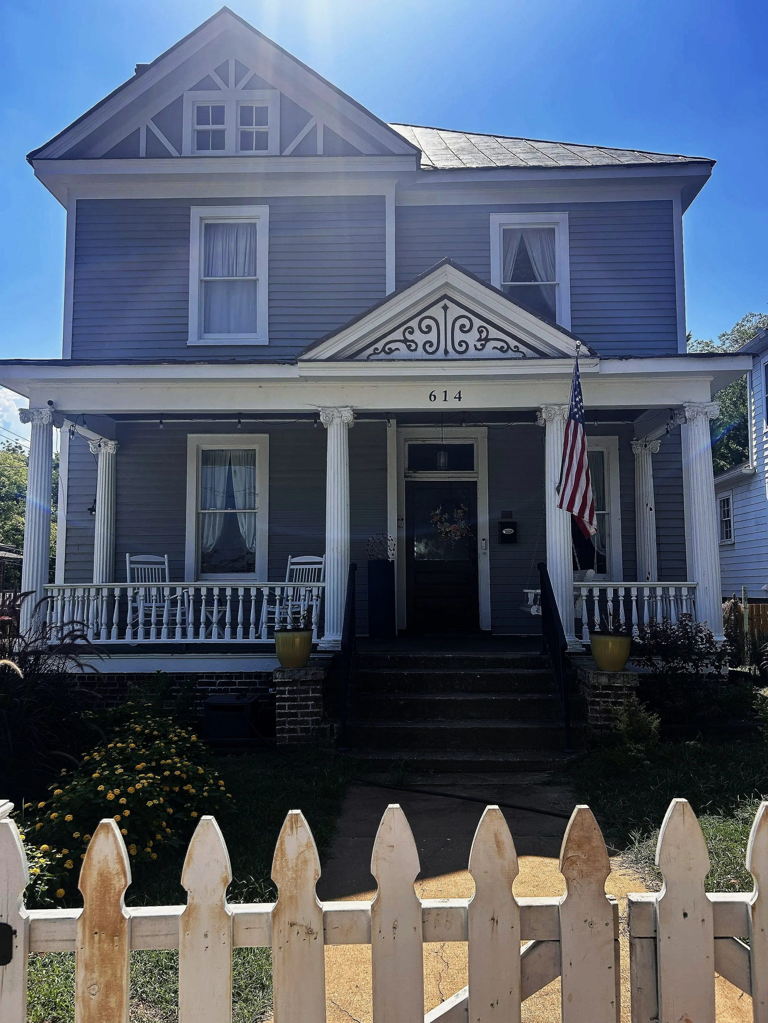 A two-story blue house with white trim, featuring a front porch with white columns, a decorative gable above the entrance, and an American flag hanging beside the door. The house has several windows with curtains and a small garden with yellow flowers in the front yard. A white picket fence encloses the yard, and sunlight creates a glow around the house.