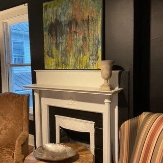 A living room with a white fireplace, colorful abstract painting above it, a beige armchair, and a striped upholstered chair.