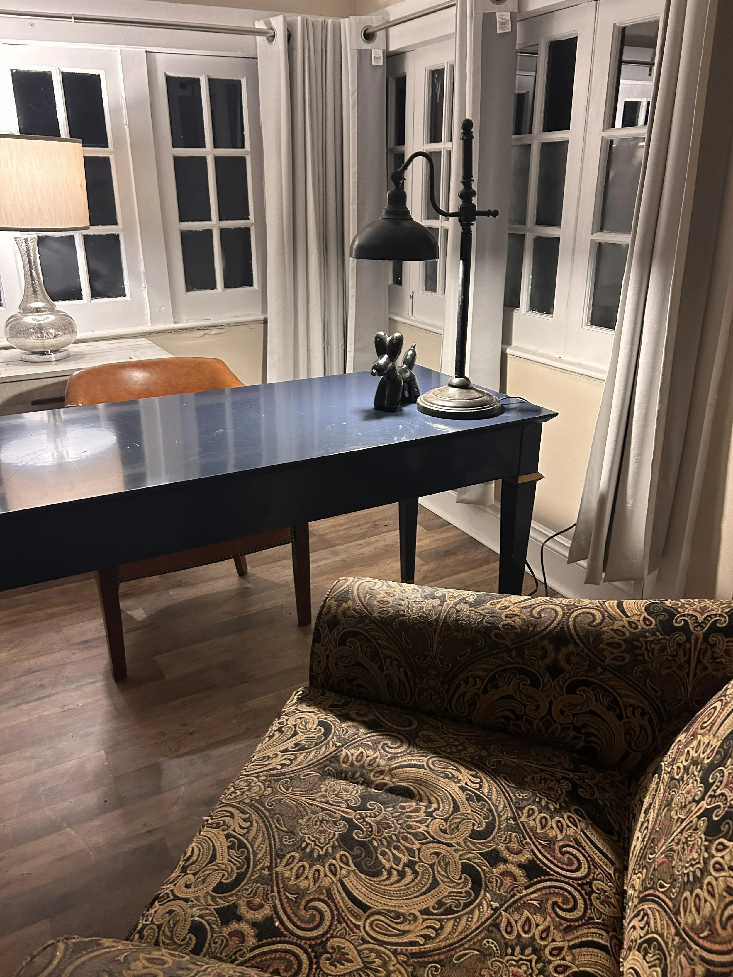 Cozy living room with a paisley pattern sofa, a dark wooden table, a lamp, and multiple windows with white curtains.