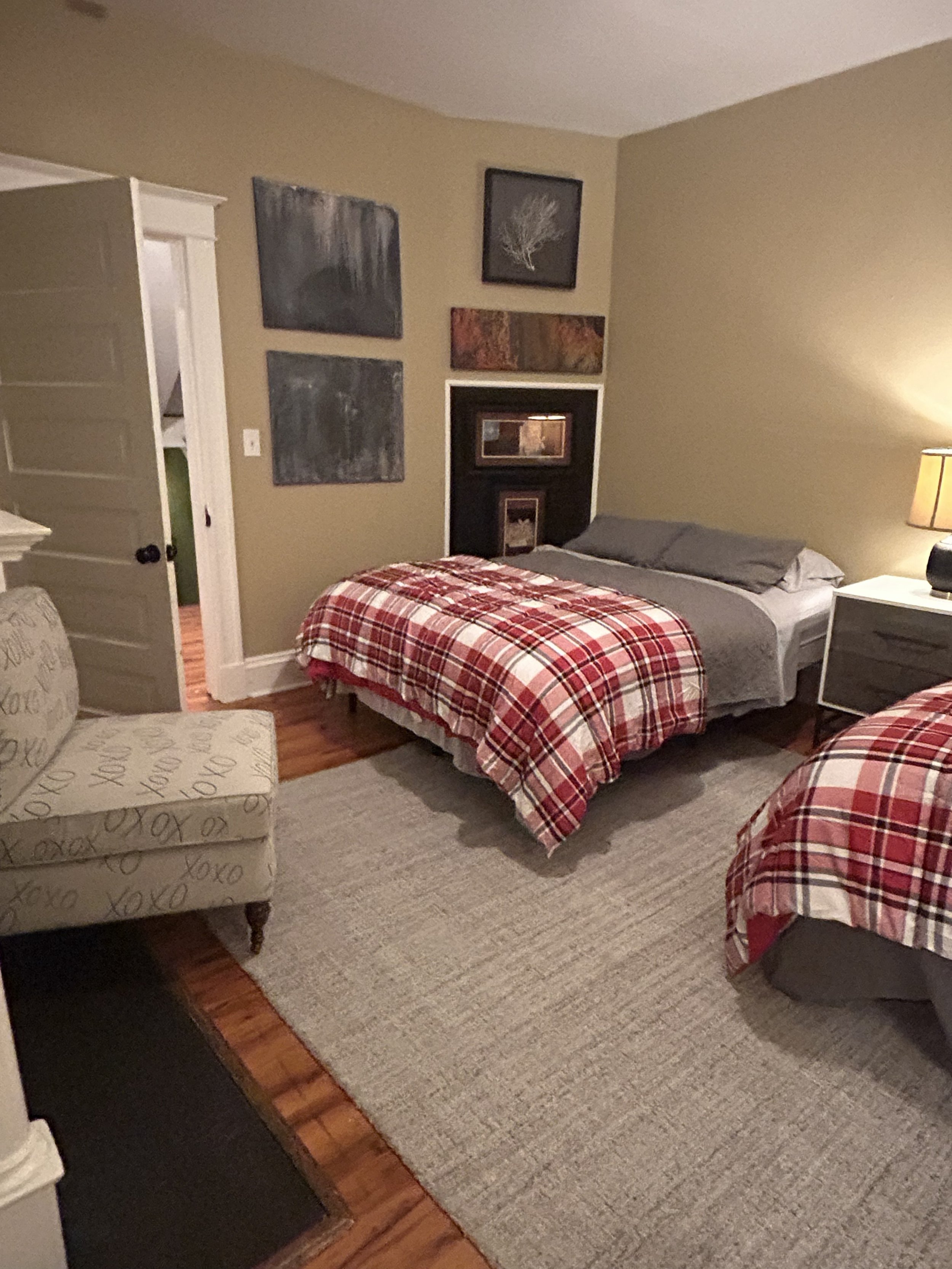 A cozy bedroom with a bed covered in a red plaid comforter, a matching plaid armchair, and beige walls decorated with several abstract paintings. There's a white nightstand with a lamp on the right, and a beige rug on wooden flooring.