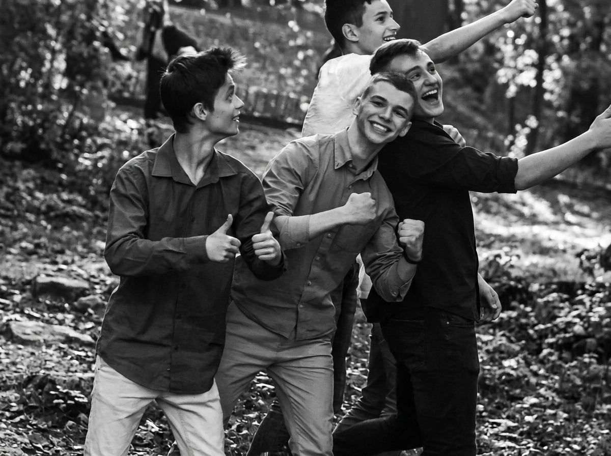 A group of four smiling friends enjoying a hike in a wooded area, some showing thumbs up and others raising their arms in excitement.