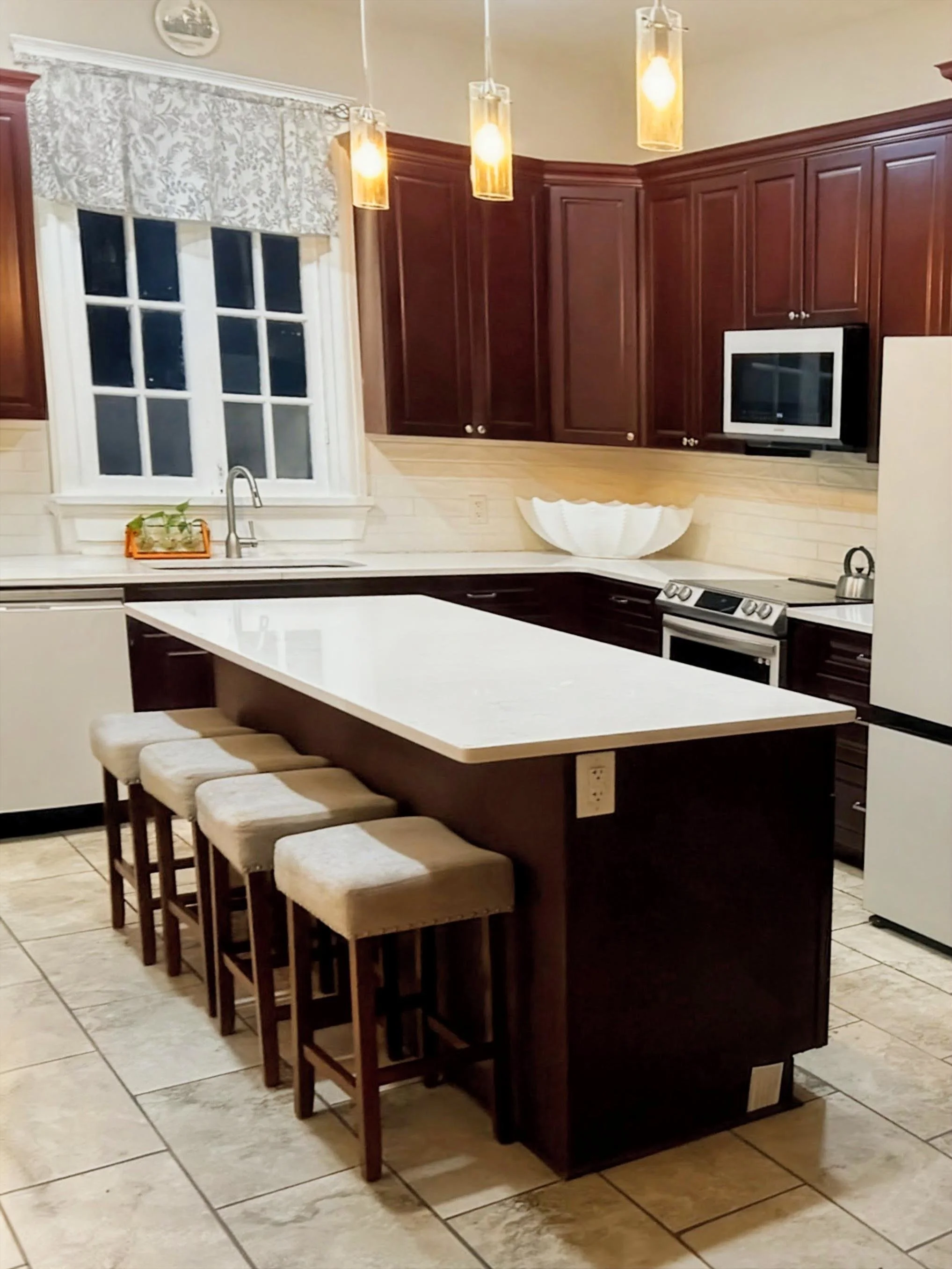 A modern kitchen with dark wood cabinets, cream-colored tile backsplash, a white countertop, a window with a white floral valance, three hanging pendant lights, a stainless steel sink with a faucet, a small potted plant on a tray, a white decorative bowl, a stainless steel oven, and beige tiled flooring.