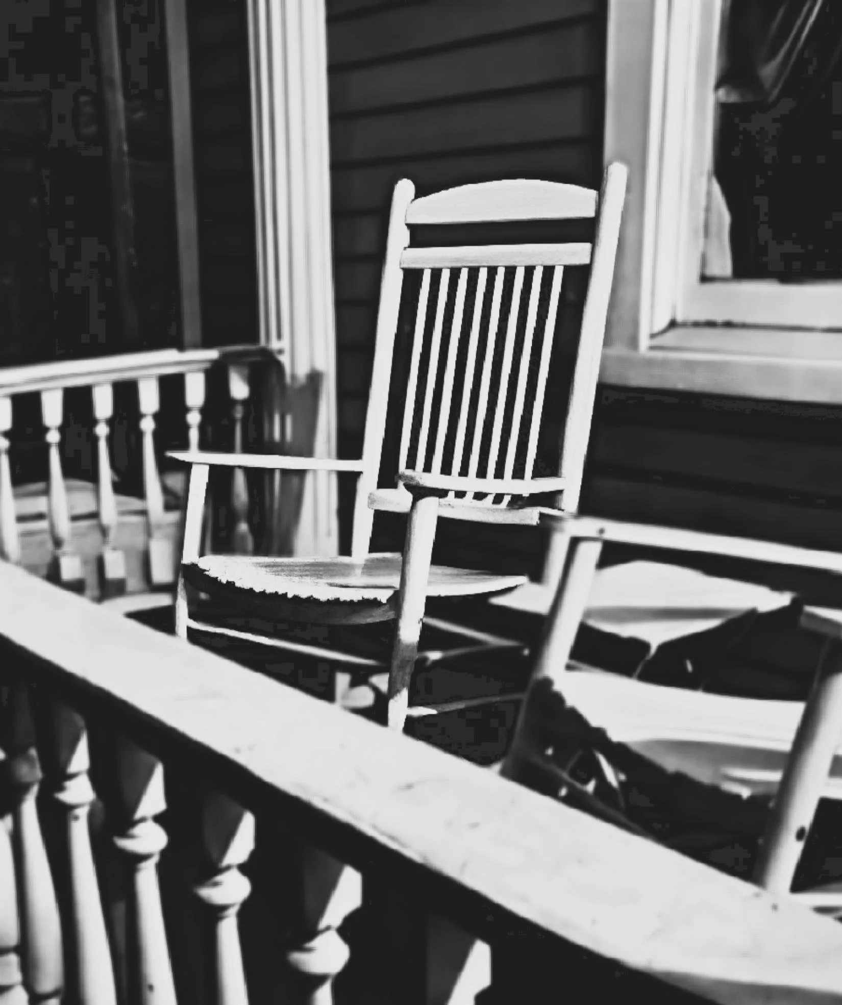 Black and white photo of wooden chairs arranged on a porch, with a window and siding in the background.