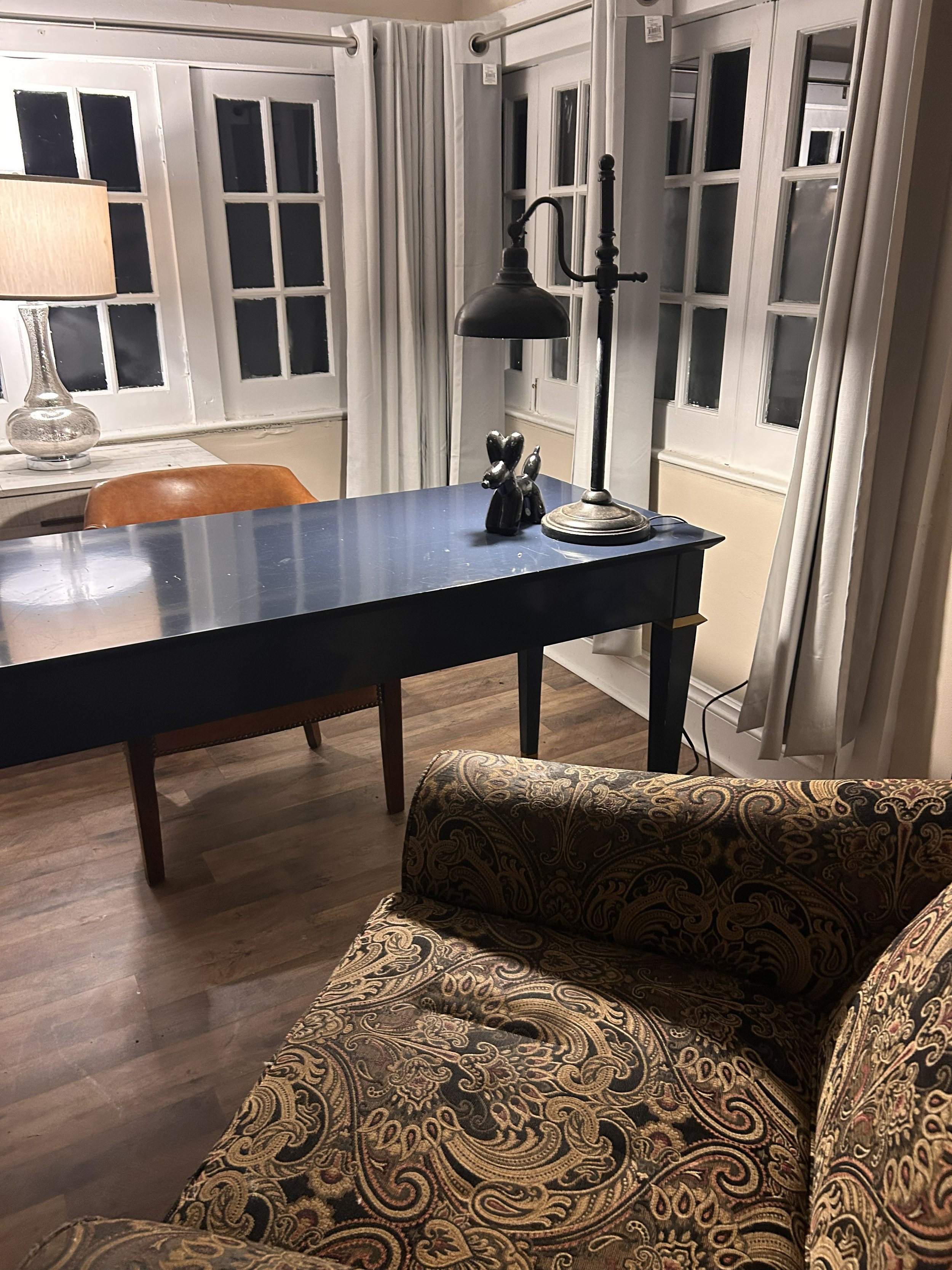 A cozy living room corner with a paisley patterned armchair, two beige curtains hanging by a window, a black table with a lamp and decorative sculptures, and a desk with a brown leather chair, a table lamp, and a large window with multiple panes.