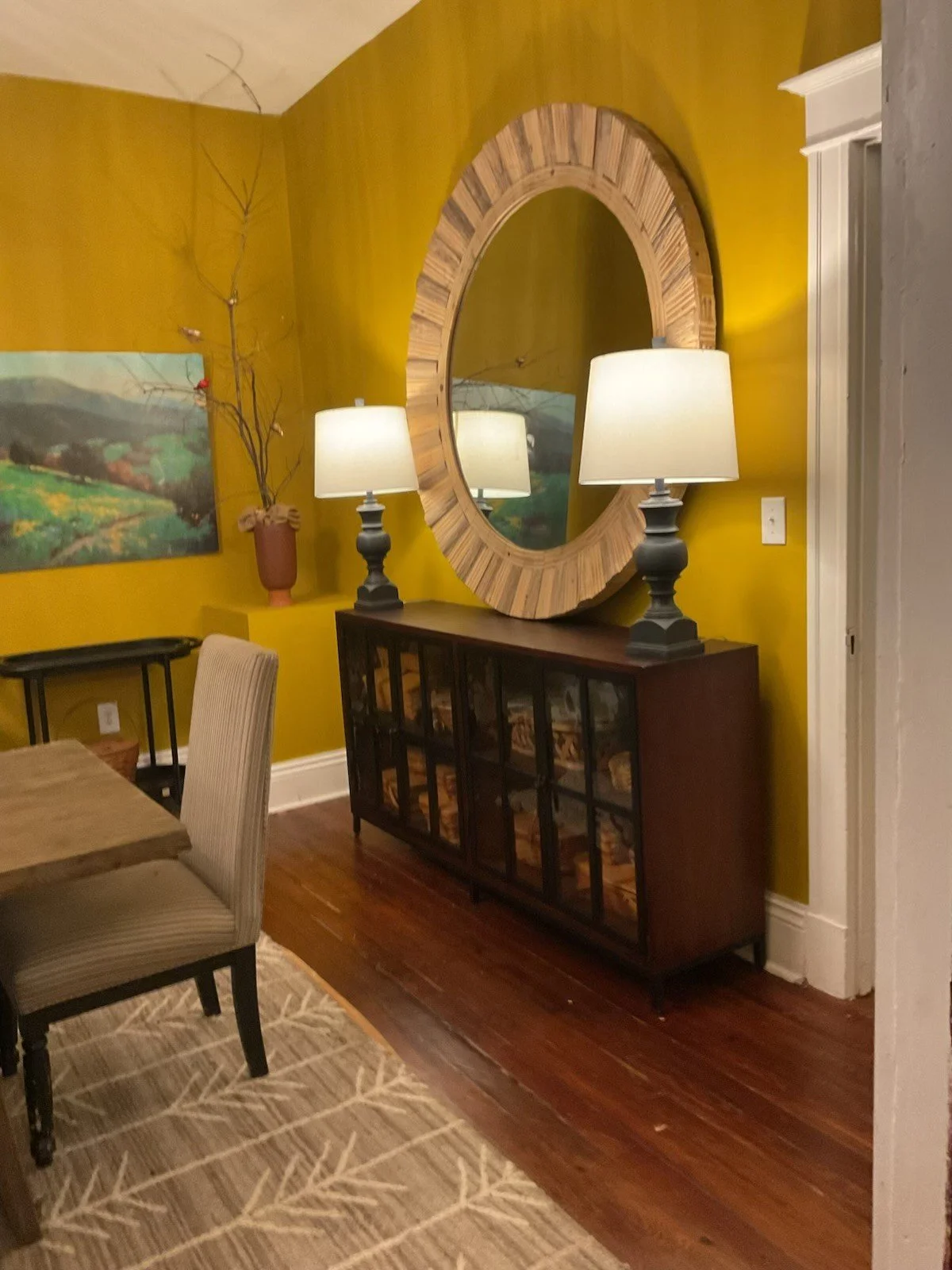 A living room corner with a yellow wall, a large round wooden mirror, two black lamps with white shades on a wooden sideboard, a beige upholstered chair, a small black table, a colorful landscape painting, a decorative branches arrangement in a red vase, and a beige rug with a leaf pattern on a hardwood floor.