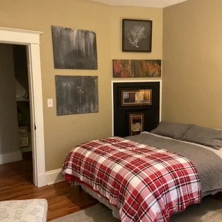 A bedroom with a bed covered in a red plaid blanket, gray pillows, and a black headboard. The wall behind has four framed art pieces in various sizes, and a door to another room is partially open.