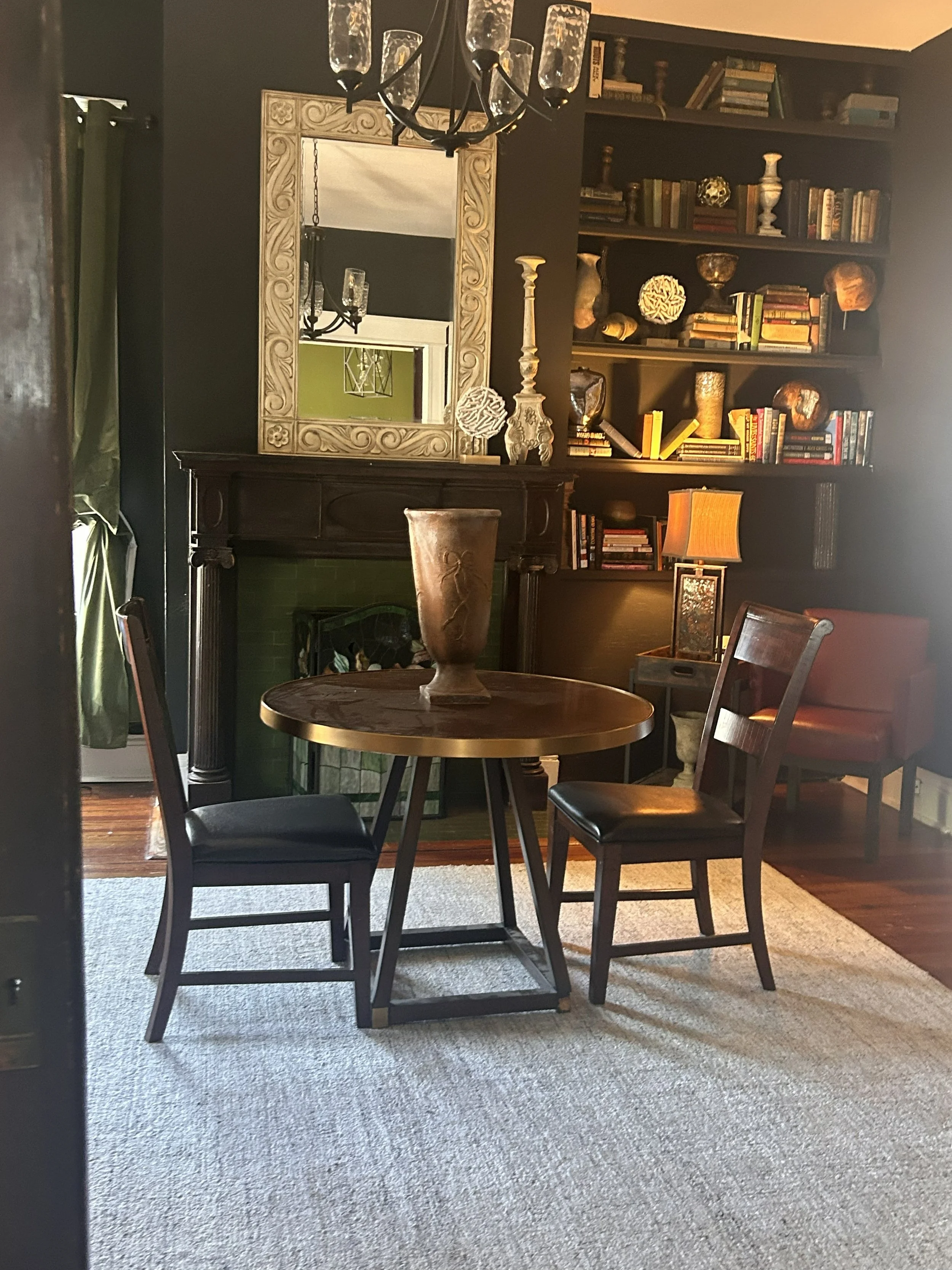 Living room with a round wooden table, four chairs, a fireplace, a large mirror, a bookshelf filled with books and decorative items, a table lamp, and a beige carpet.