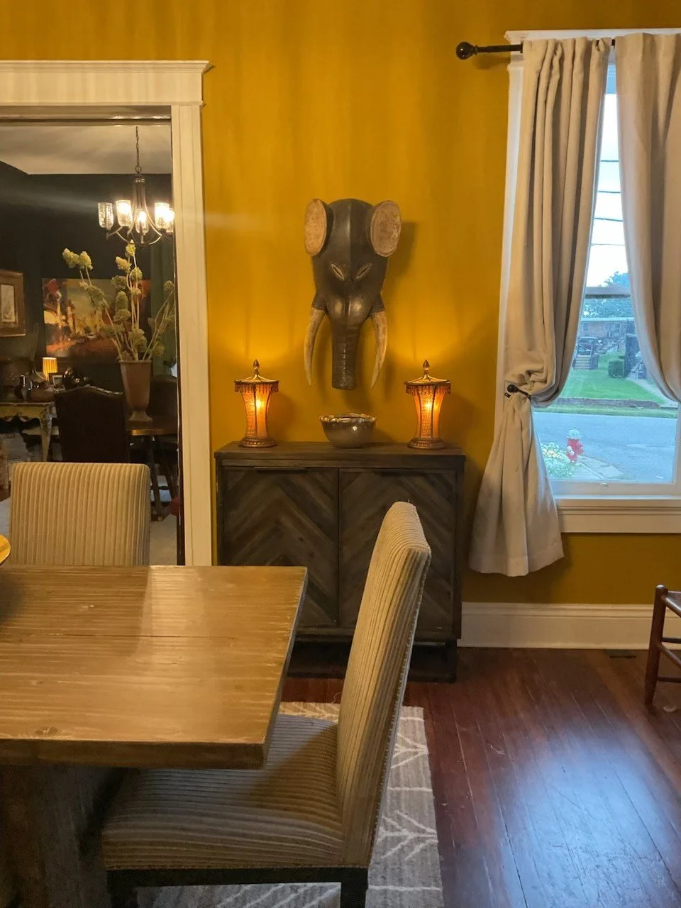 A dining room corner with a yellow wall, a window with white curtains, a wooden sideboard, a tribal-style mask on the wall, and two lamps with orange shades on the sideboard.
