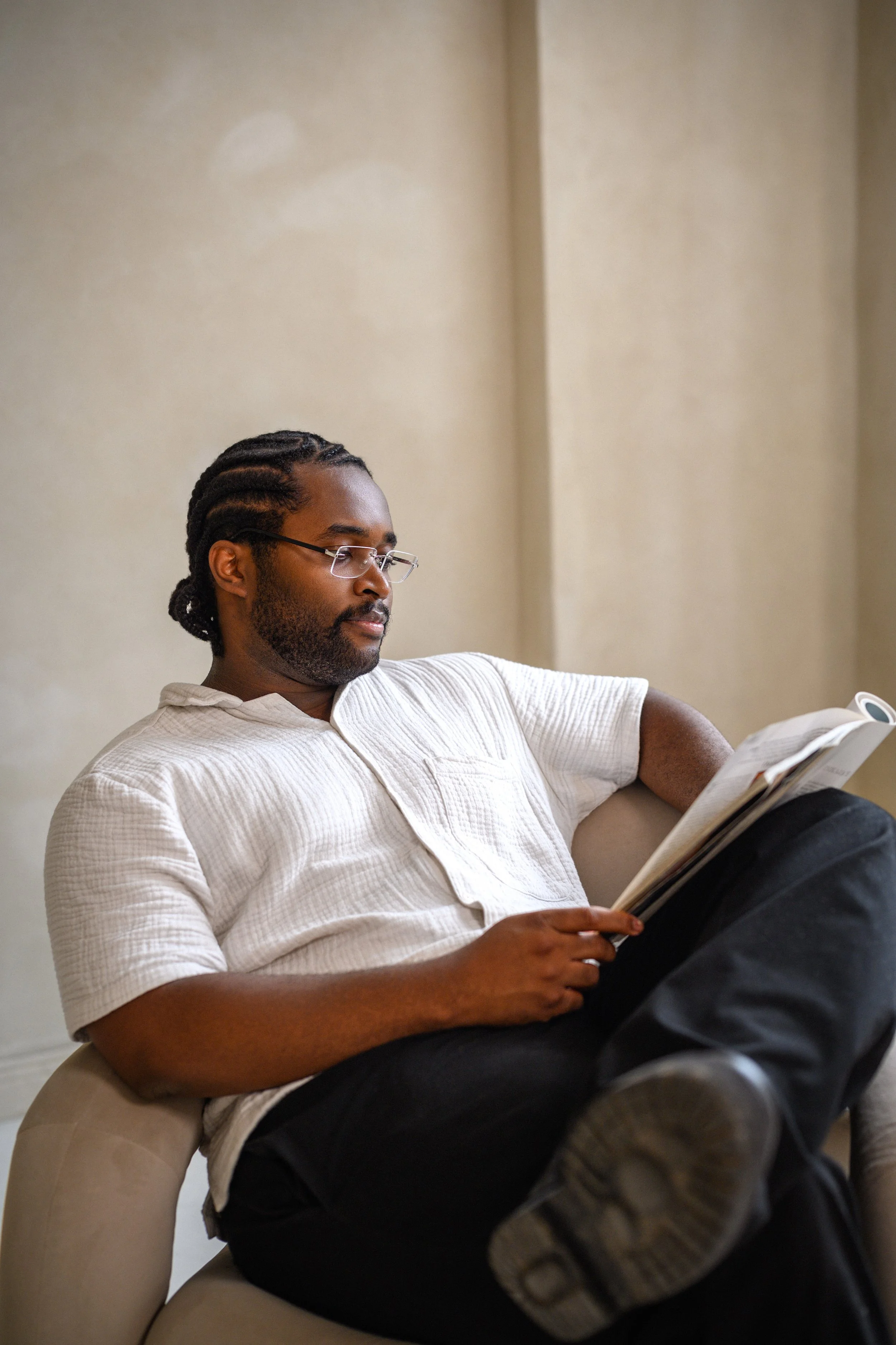 A man with glasses and dreadlocks sits on a beige chair, reading a magazine in a room with beige walls.