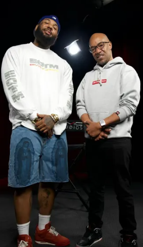 Two men standing in a studio, one wearing a white sweatshirt, denim shorts, and red sneakers, the other in a hoodie and glasses, with studio lighting and equipment in the background.