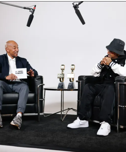An interview setting with two men seated in black armchairs, microphones hanging overhead, and a small table with two awards or trophies between them.