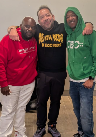 Three men standing together, smiling, with their arms around each other in an indoor setting. The man on the left wears a red hoodie, the man in the middle wears a black t-shirt, and the man on the right wears a green hoodie.