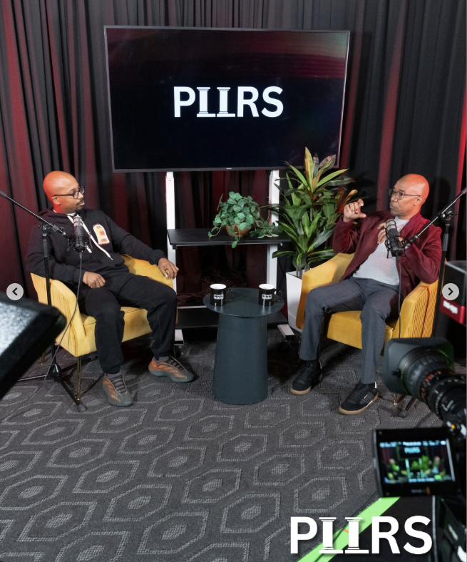 Two men recording a podcast or interview in a studio with black curtains, yellow chairs, and a large screen displaying the word 'PLLRS'.