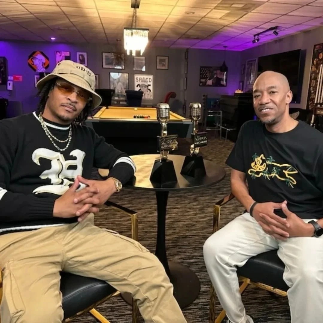 Two men sitting at a round table with a yellow pool table behind them, decorated with vintage pictures, sculptures, and neon lights.