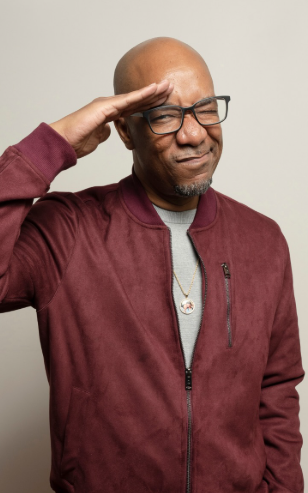 Older man with glasses saluting or shielding his eyes, wearing a maroon jacket and gray shirt, standing against a plain light background.
