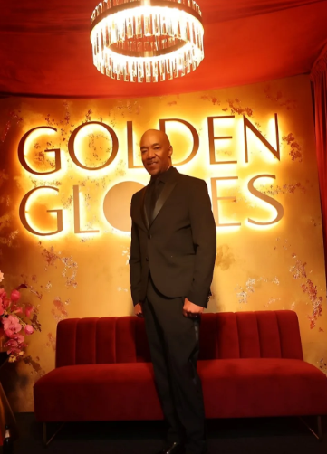 Man in a black suit standing in front of a glowing sign that says "Golden Glories" with a red couch and a chandelier overhead.