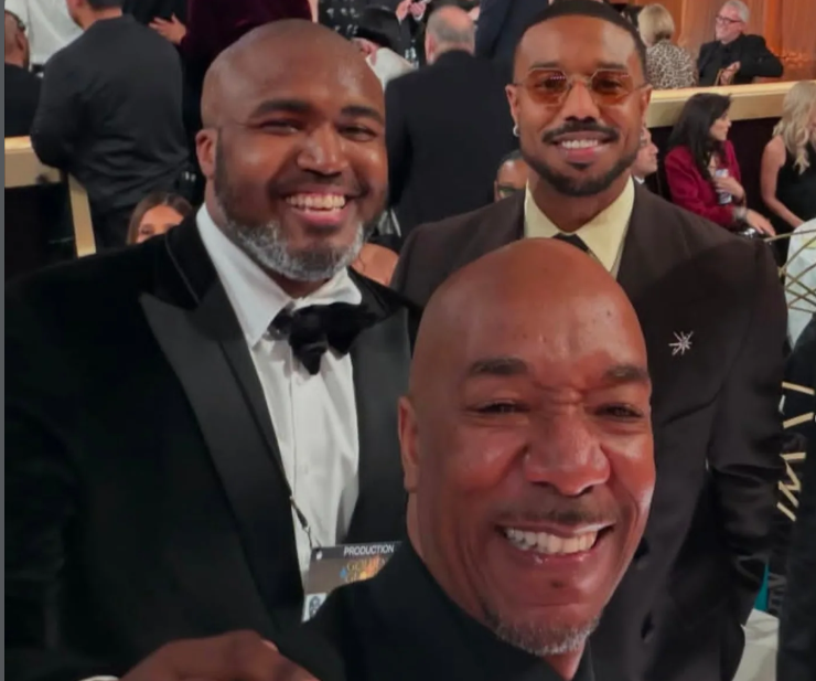 Three men dressed in formal attire smiling at a social event. The man in the foreground is taking a selfie, the man on the left is wearing a tuxedo with a bow tie, and the man on the right is wearing glasses and a suit.