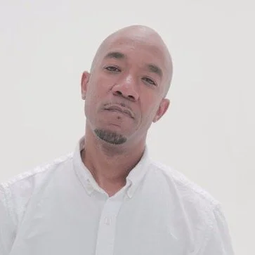 A bald man with a goatee wearing a white collared shirt, standing against a plain light-colored background.