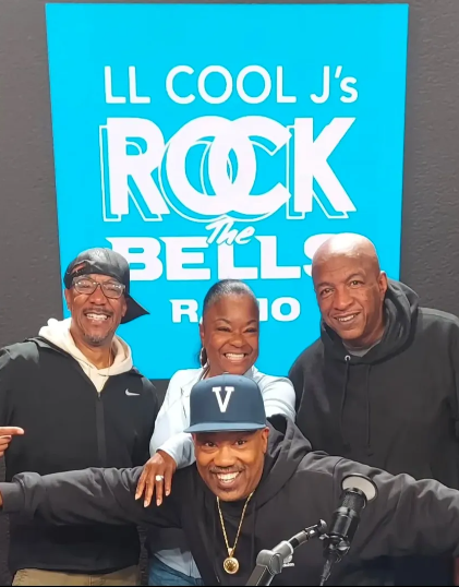 Four people smiling in front of a bright blue sign reading 'LL COOL J's ROCK THE BELLS RADIO.'