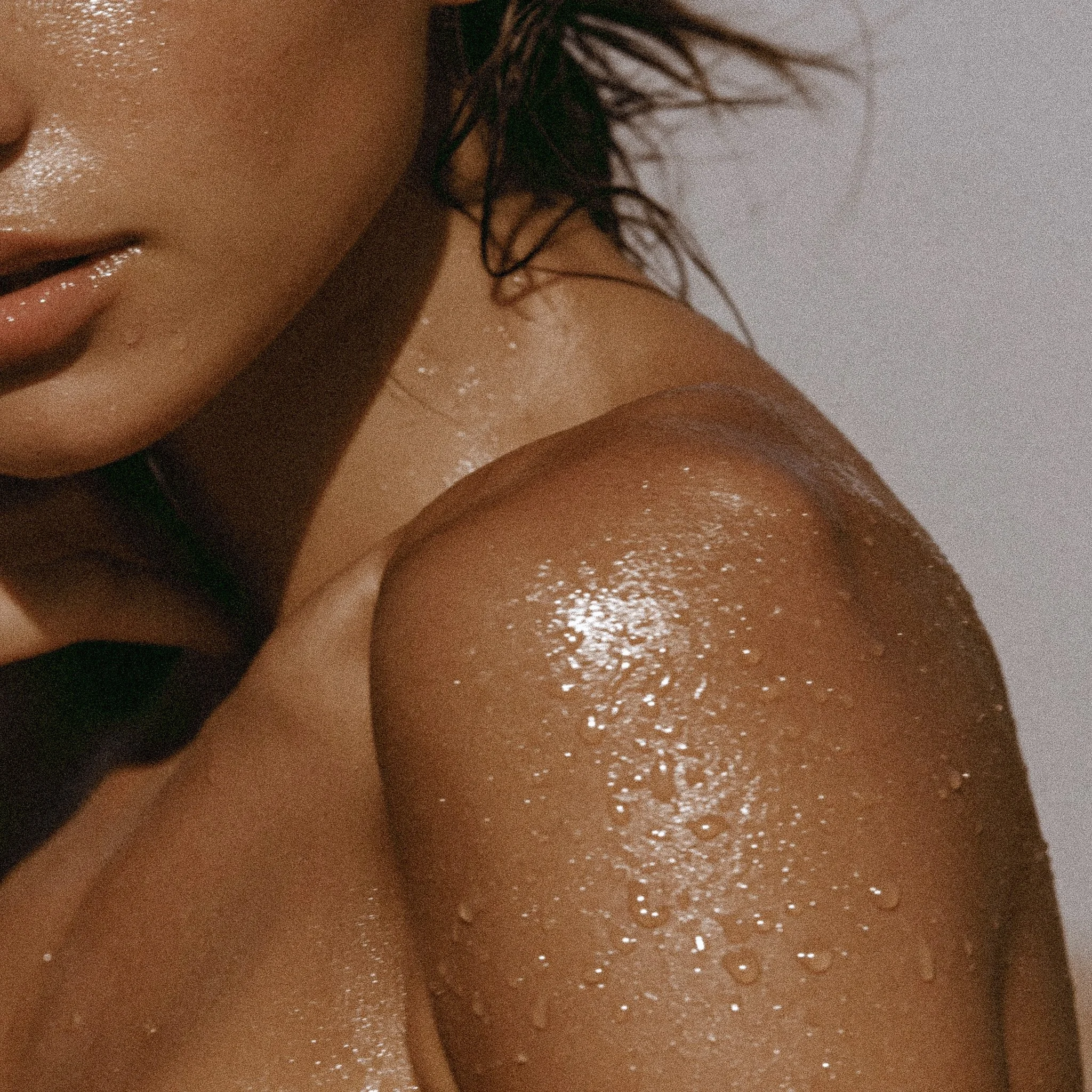 Close-up photo of a woman's face and shoulder, with wet skin, dark curly hair, and glitter on her skin, using a shower or bath.