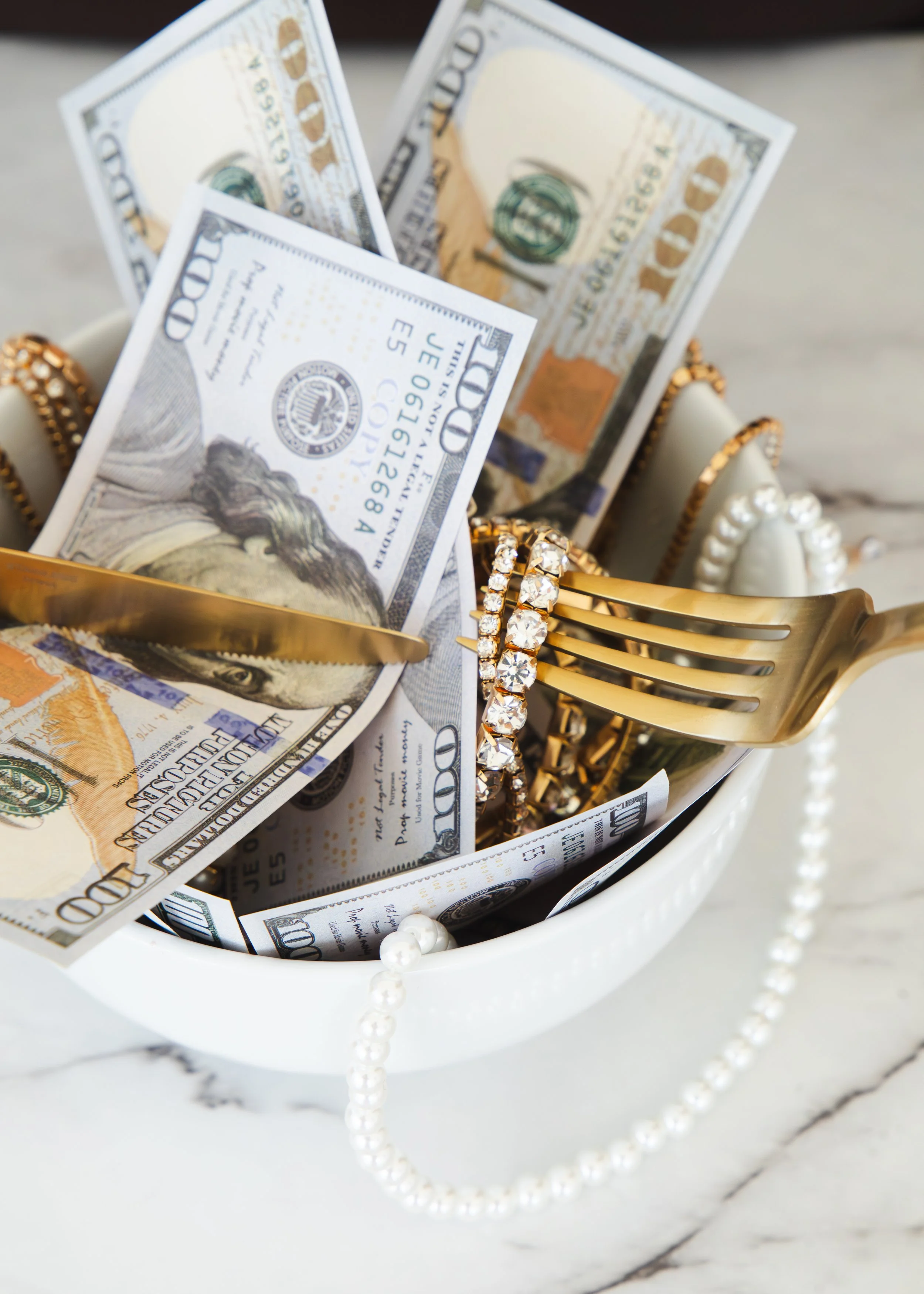 A white bowl filled with crumpled 100-dollar bills, gold jewelry with clear gemstones, a gold fork, and a string of pearls, on a marble surface.