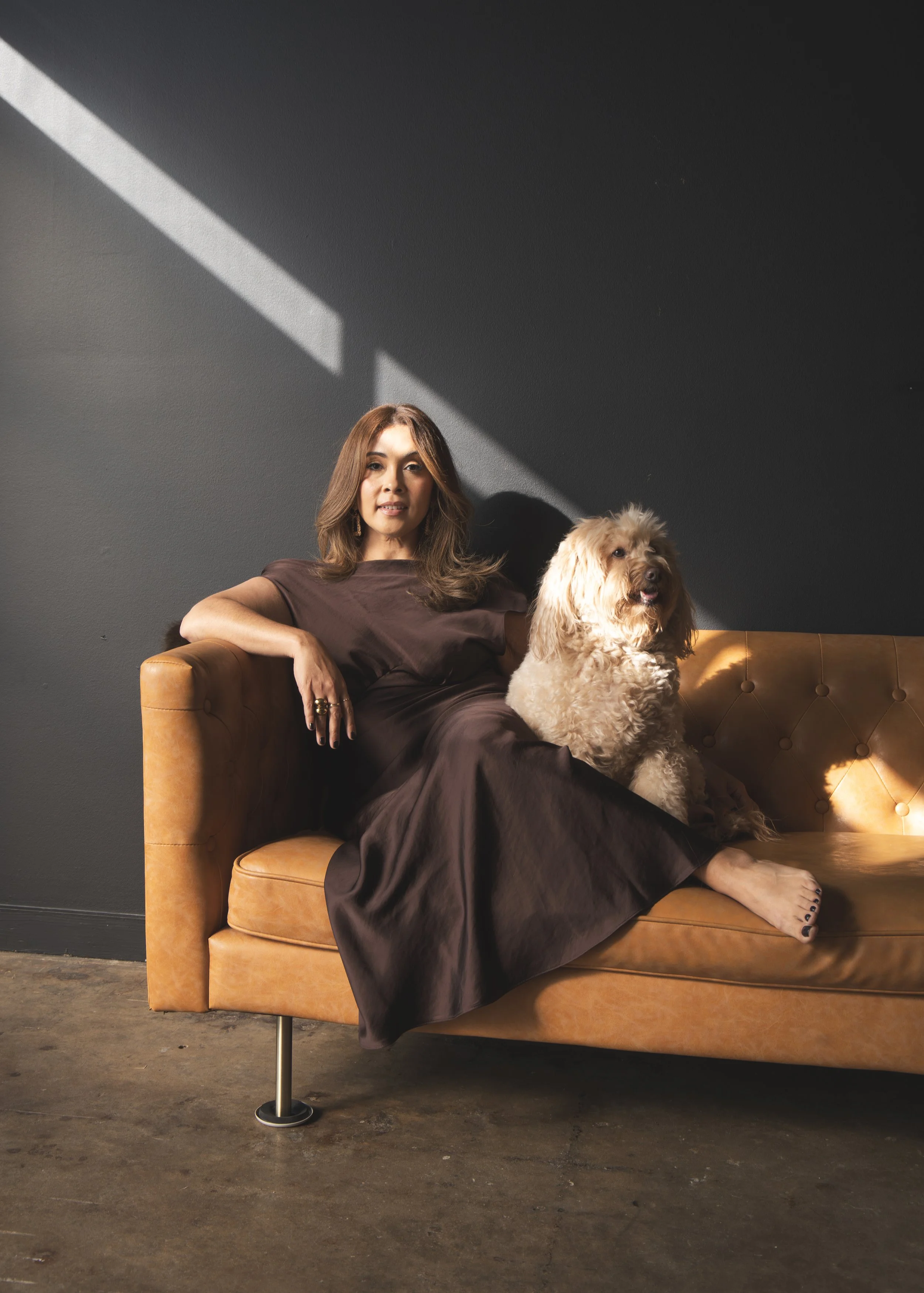 A woman with brown hair wearing a dark brown dress, sitting on a tan leather sofa with a large, curly-haired dog. They are positioned against a dark gray wall with a diagonal sunlight shadow.