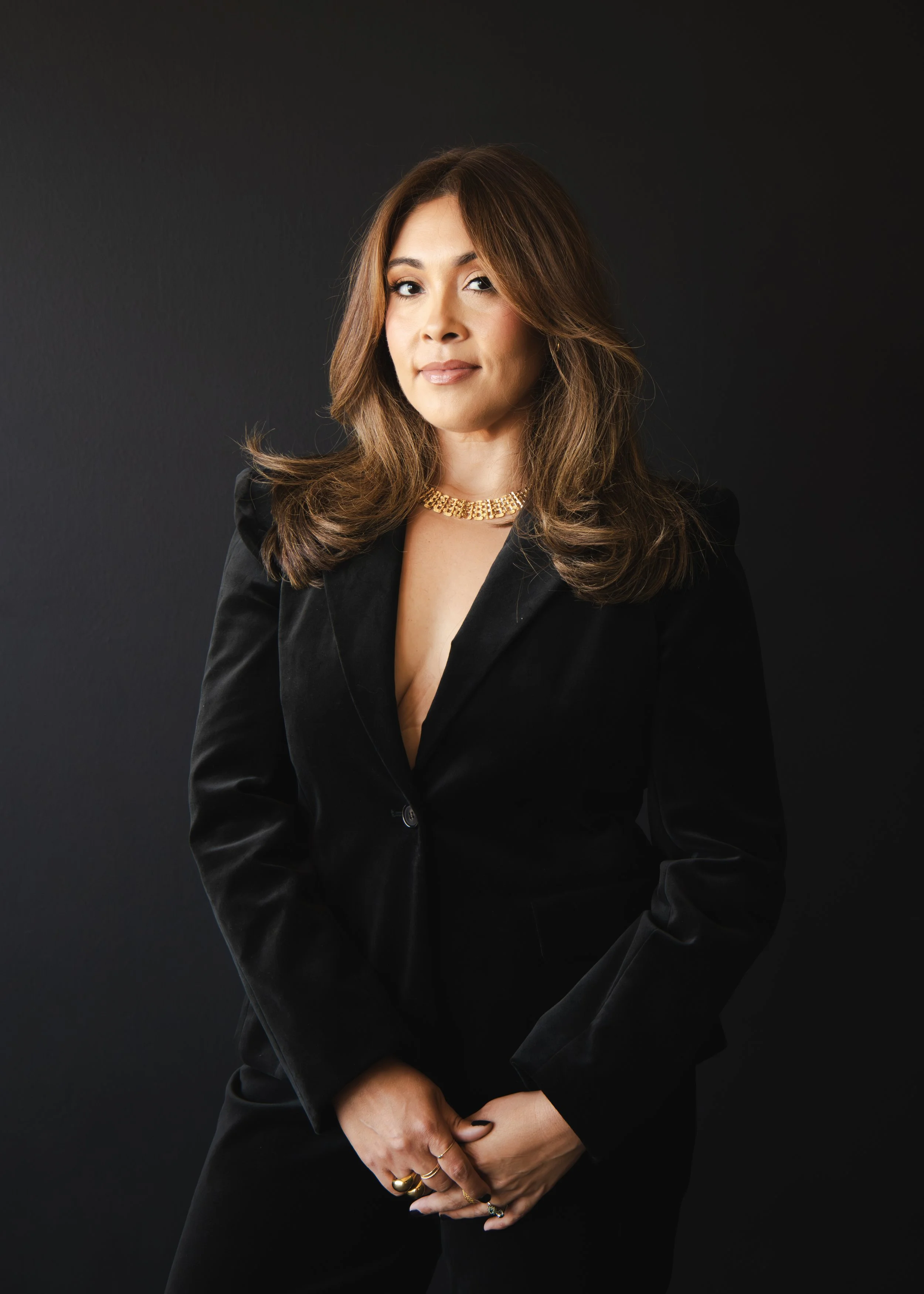 A woman with brown hair wearing a black blazer, gold necklace, and rings posing against a dark background.