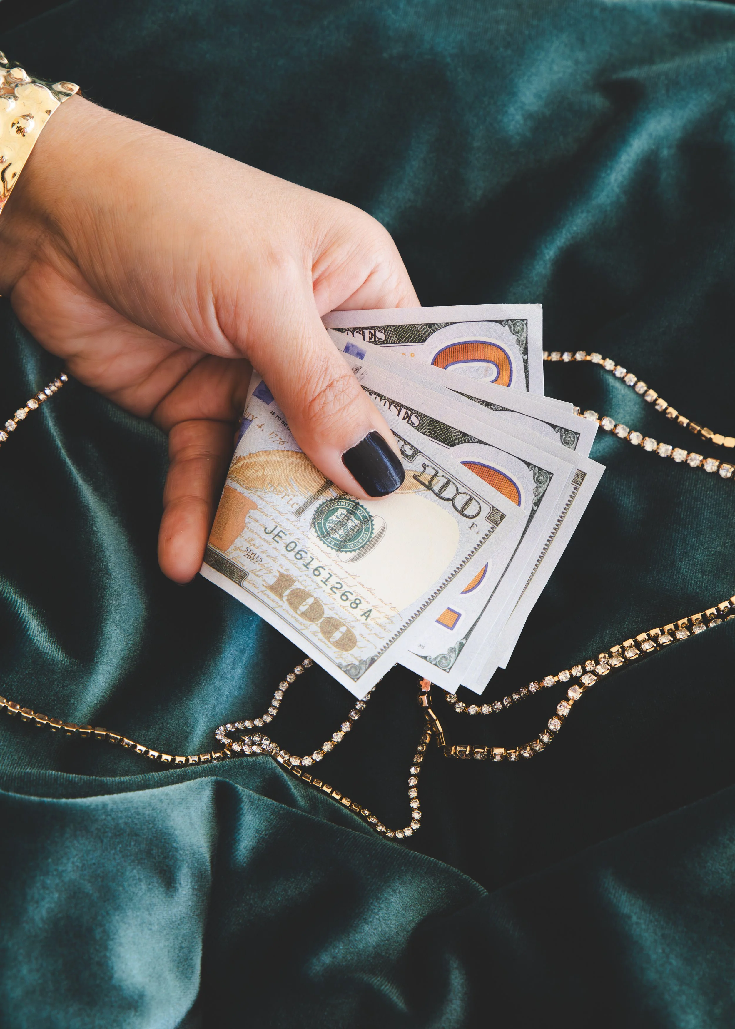 A hand with black painted nails holding three 100-dollar bills, resting on dark green silky fabric with diamond and gold jewelry.