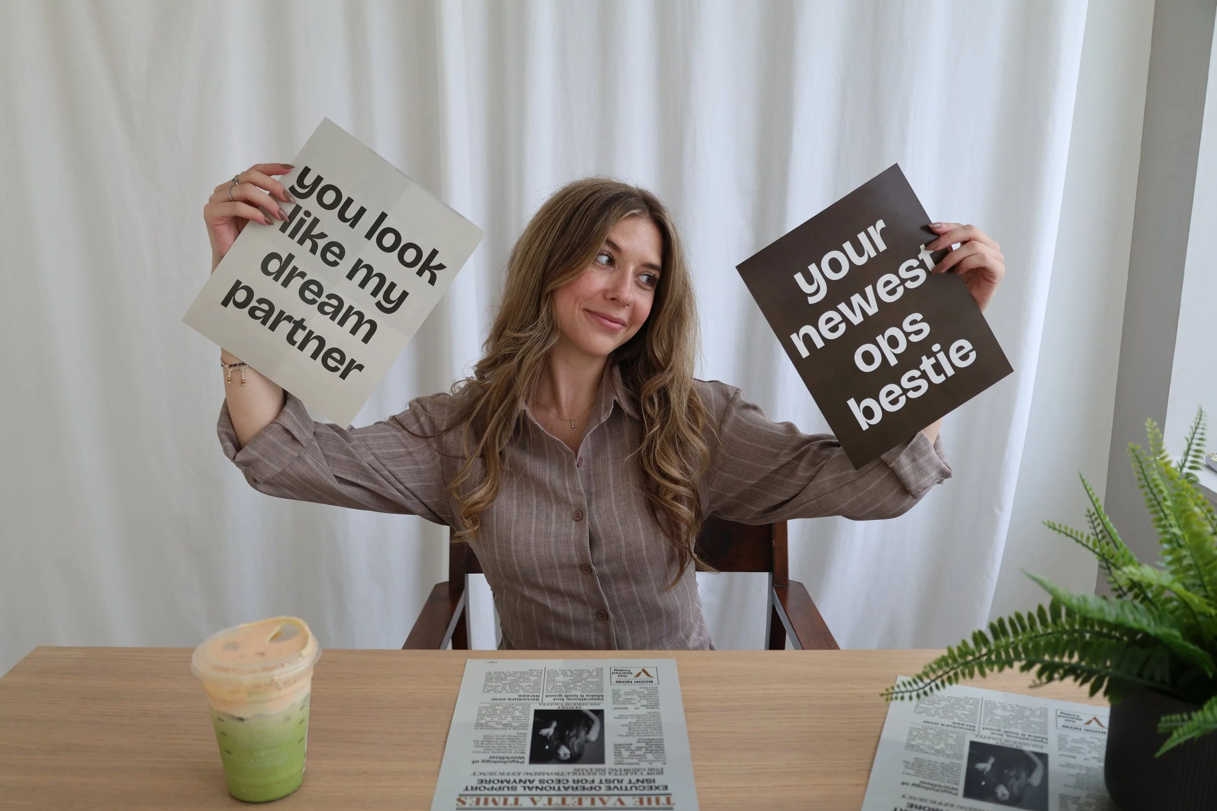 A woman with long brown hair and a checkered shirt sitting at a table holding two signs, one saying 'you look like my dream partner' and the other saying 'your newest bestie'. There is a multicolored ice cream in a cup on the table, with some papers and a plant visible.